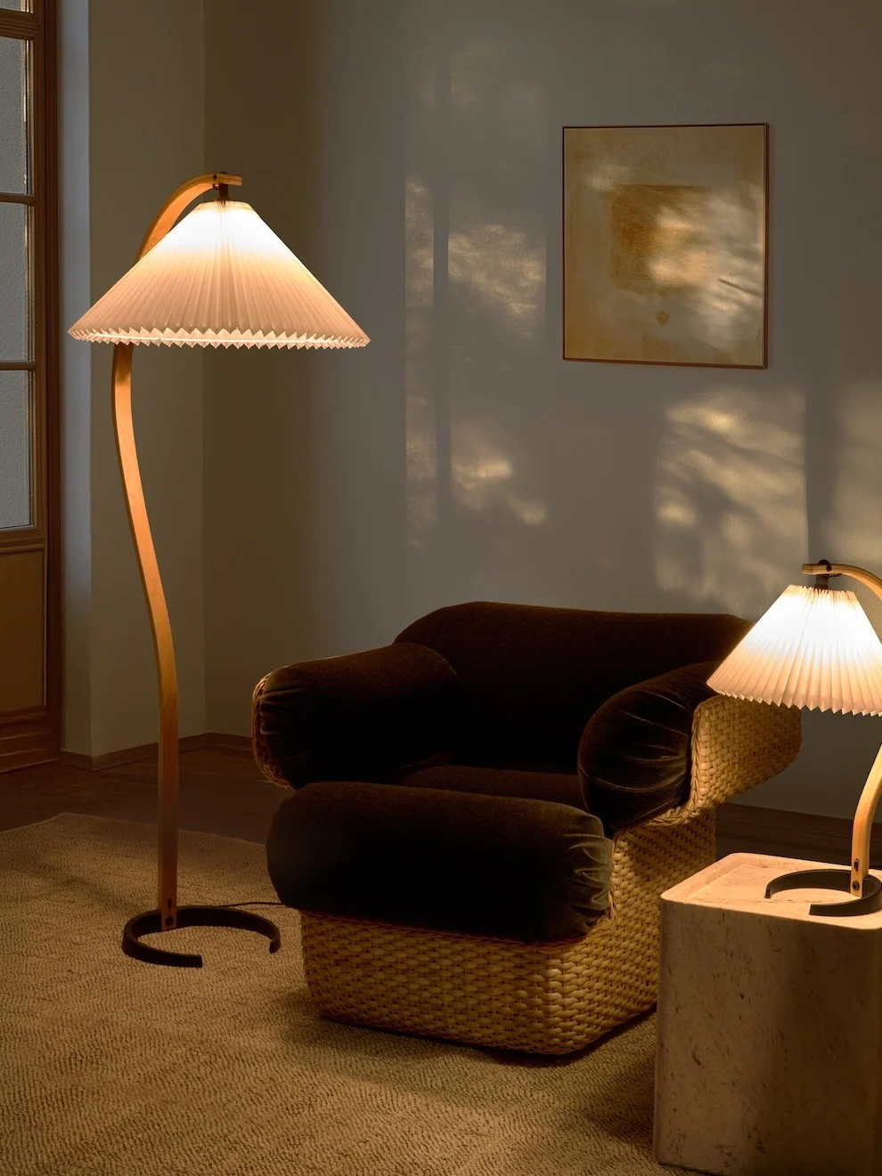 Gemütlicher Raum mit einer gebogenen Stehlampe, dunklem Sessel mit geflochtenem Untergestell und einer kleineren Tischlampe. Warmes Licht wirft Schatten.