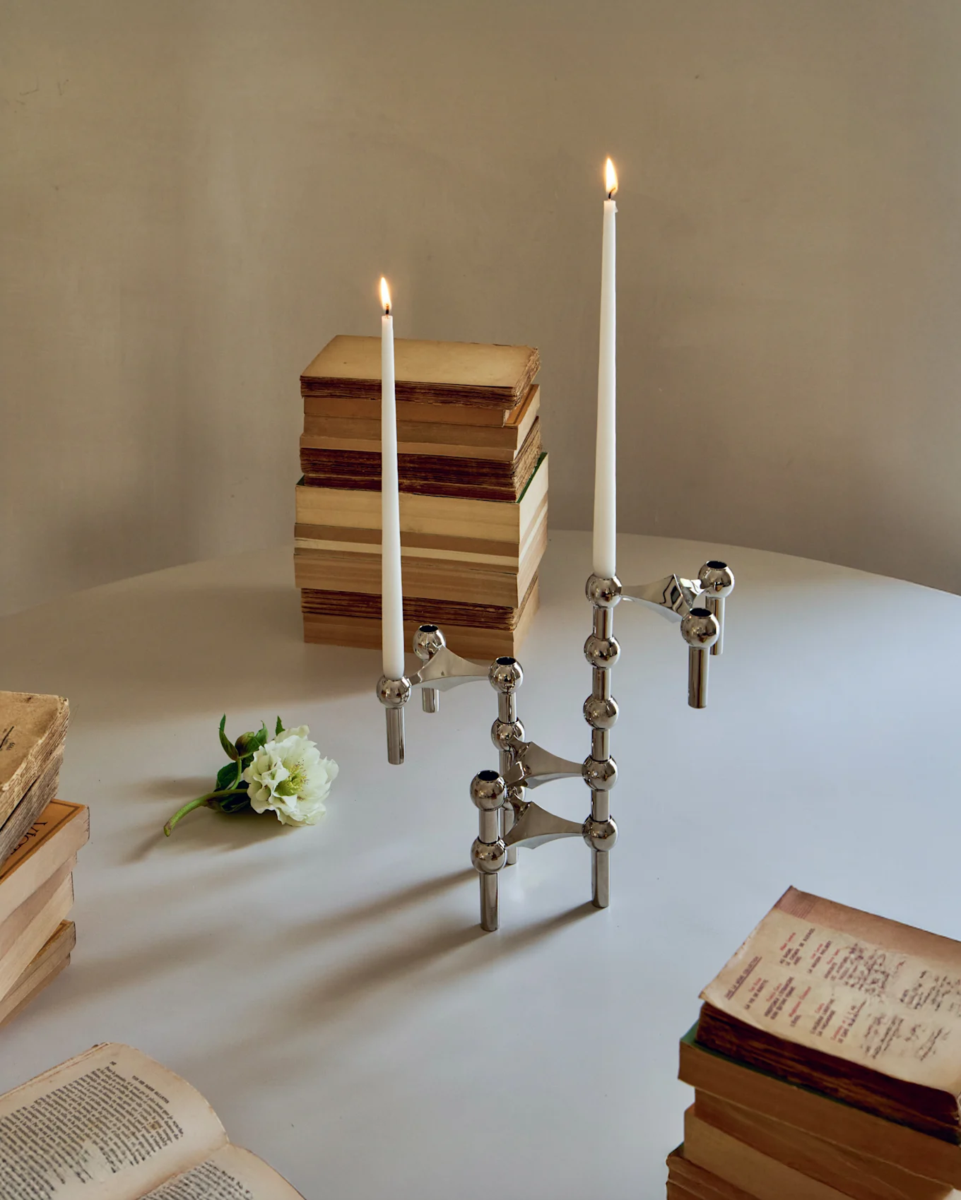 Zwei modulare Chrom-Kerzenständer mit brennenden weißen Stabkerzen, umgeben von alten Büchern und einer weißen Blume auf einem Tisch.