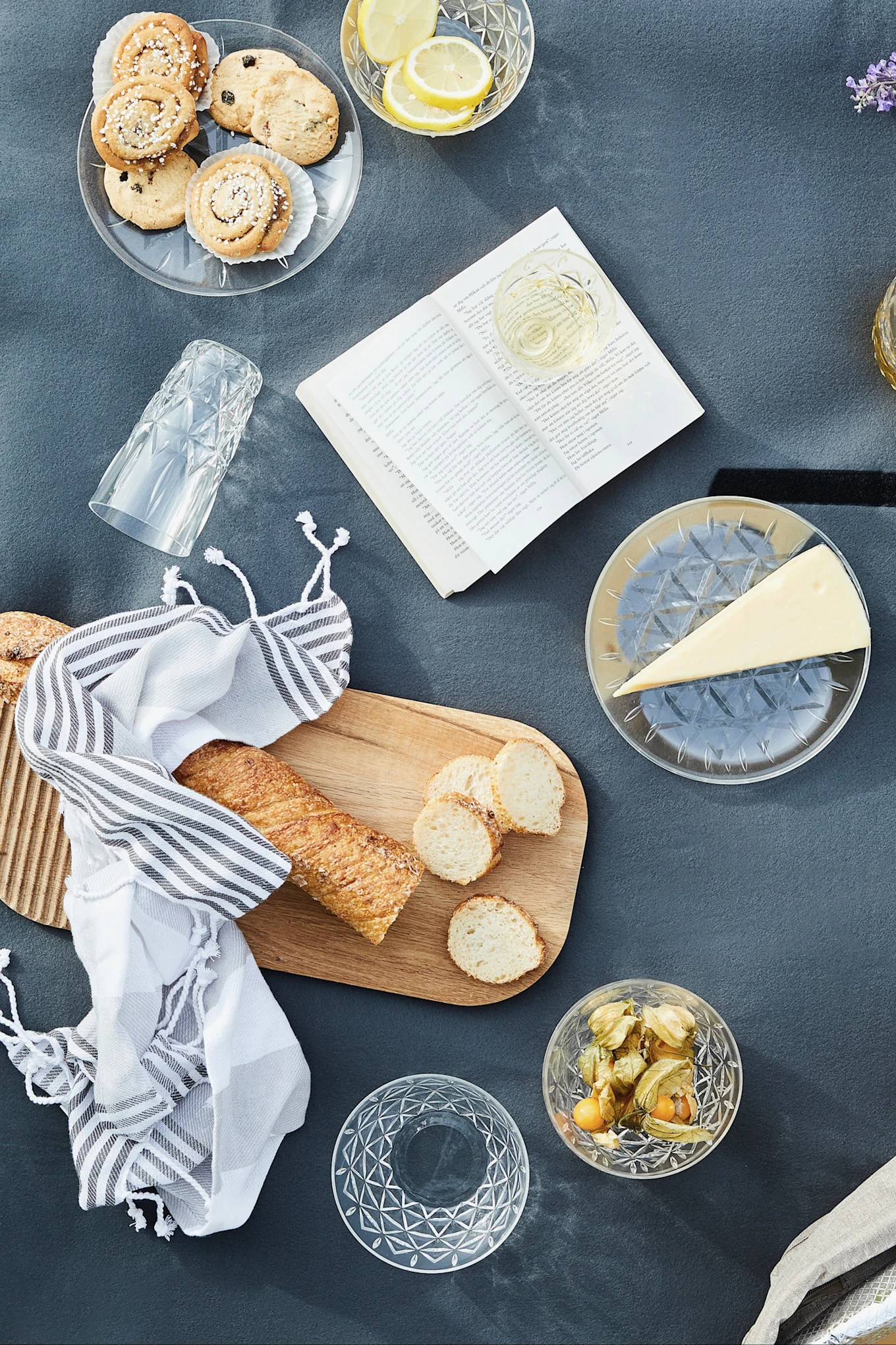 Blick von oben auf ein Picknick mit Keksen, Zitronen, Wein, Käse, Baguette, Physalis und einem offenen Buch auf einer grauen Decke.