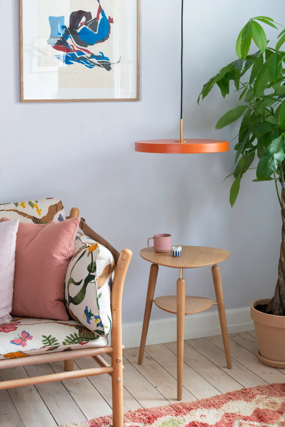 Orangetöne eignen sich perfekt für den Boho-Stil. Hier sehen Sie eine Bank mit bunten Kissen, daneben der My Spot Beistelltisch und der Asteria Pendelleuchte, beides von Umage.