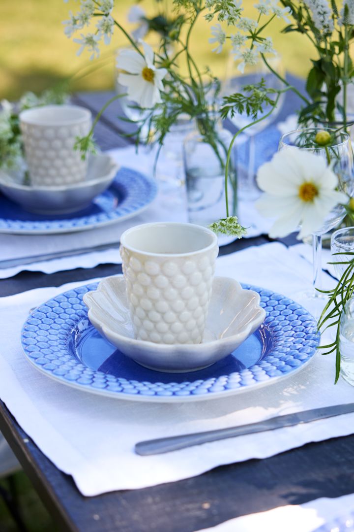 Eine Nahaufnahme eines in blau-weiß gedeckten Tisches im Freien. Ein blauer Bubbles-Teller zusammen mit einem weißen Bubbles-Becher auf einem mit Blumen geschmückten Tisch.