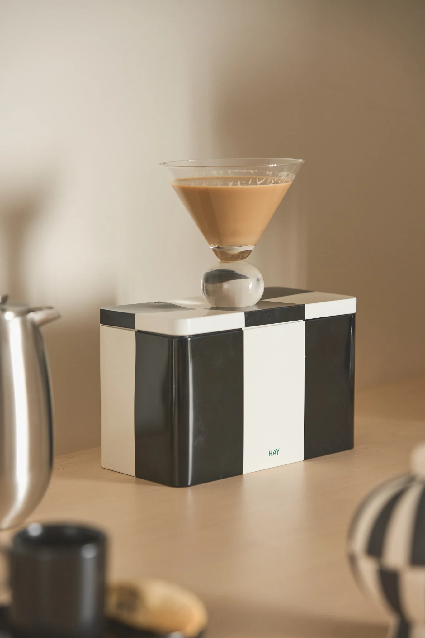 Martini-Glas mit hellbraunem Getränk und kugelförmigem Stiel auf einer schwarz-weiß karierten Box mit 'HAY'-Logo.