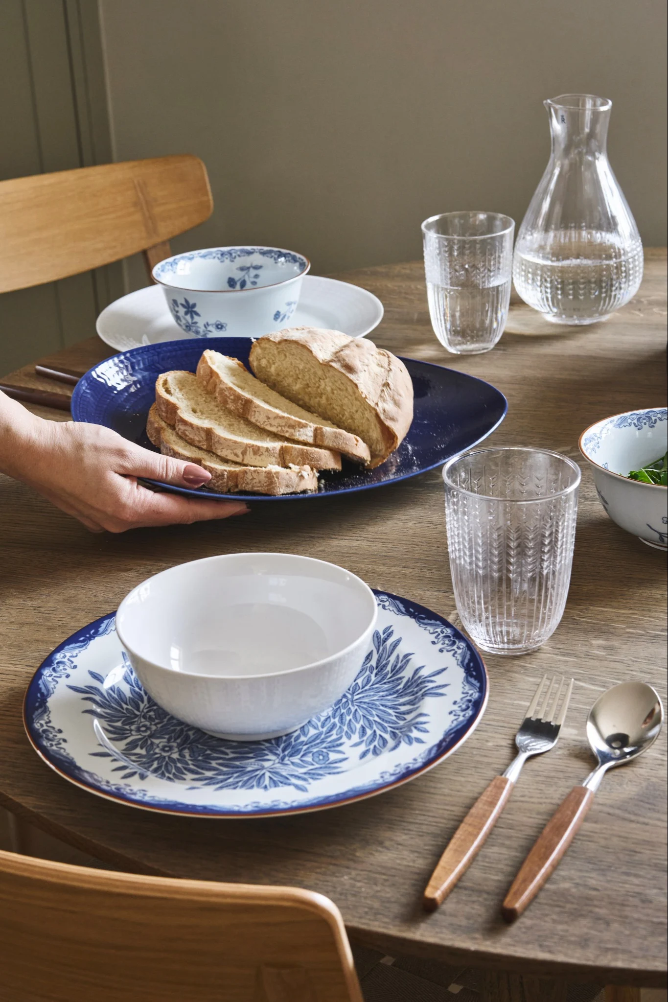 Hände platzieren eine blaue Platte mit geschnittenem Brot auf einem Tisch mit blau-weißem Geschirr, Gläsern und Besteck.