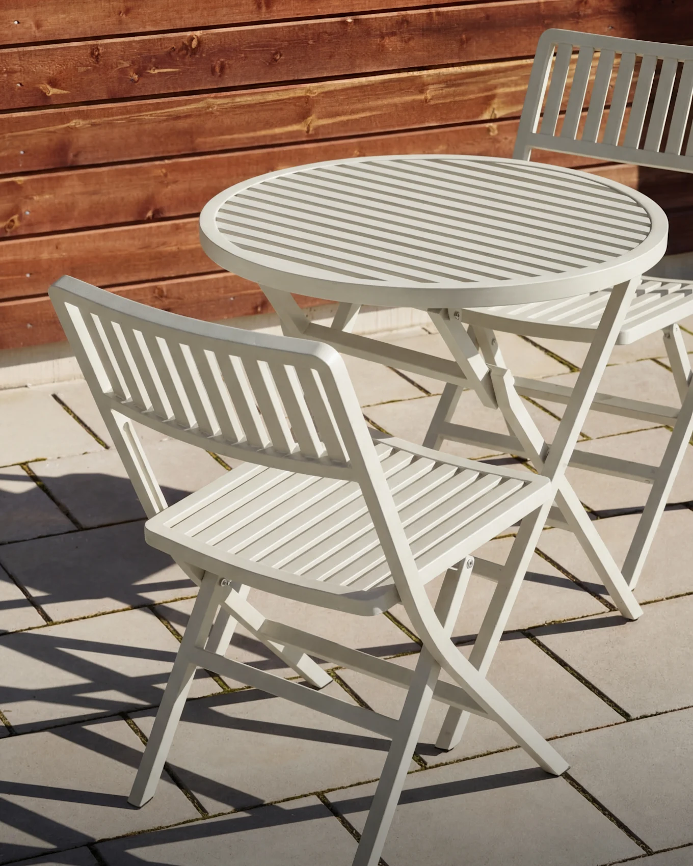 Hellgrauer Lamellen-Gartentisch mit zwei passenden Klappstühlen auf einer Terrasse.