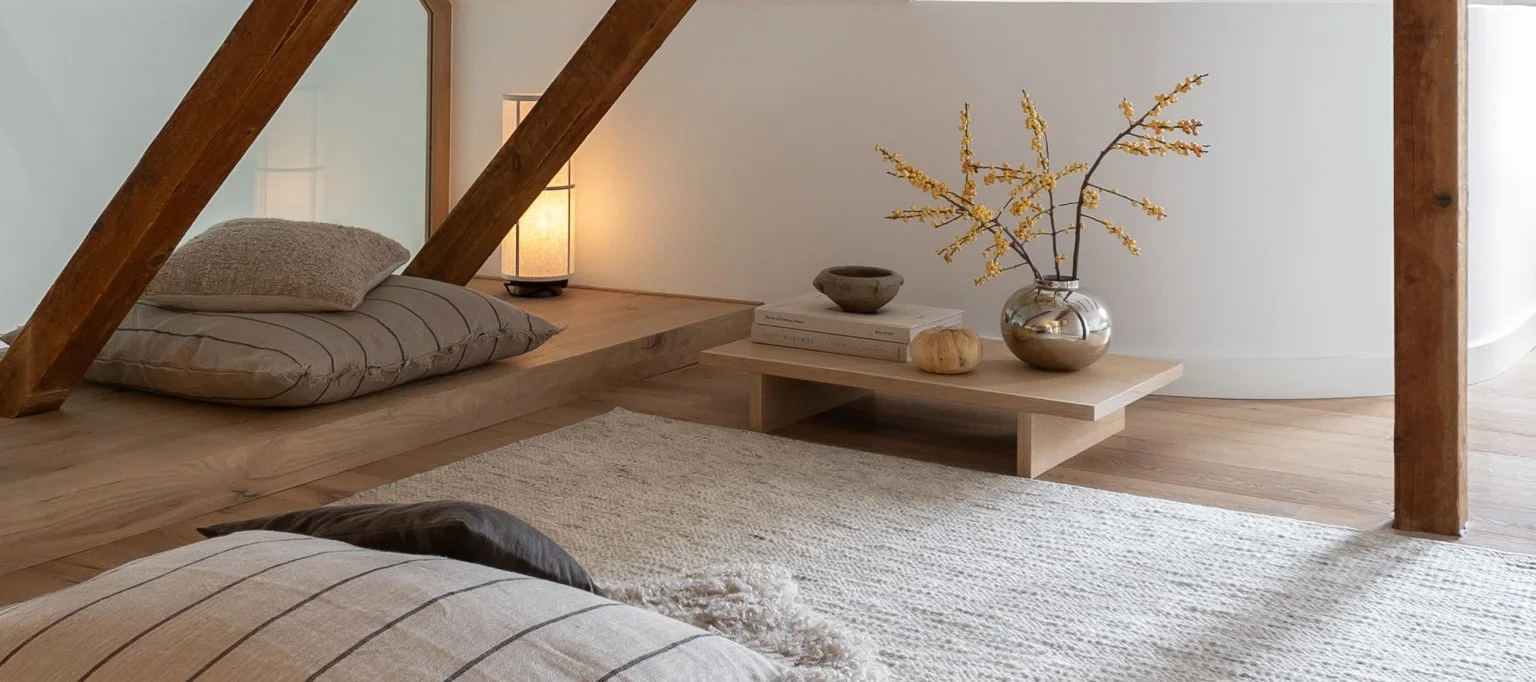 Ein gemütlicher Raum mit hellen Holzböden und weißen Wänden, mit Bodenkissen, einer leuchtenden Lampe und einem niedrigen Tisch mit einer Silbervase mit gelben Beeren.