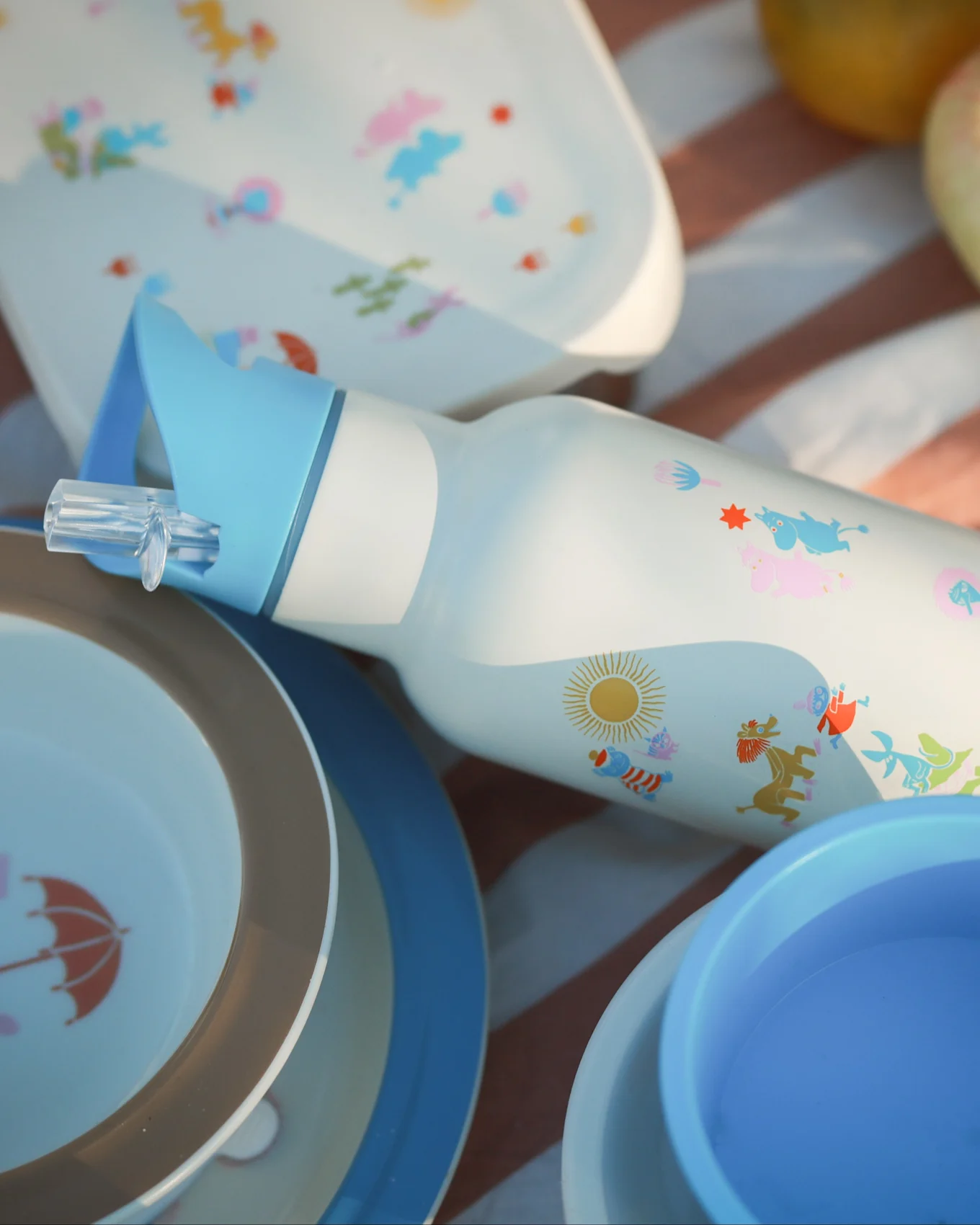Weiße Wasserflasche mit blauem Strohhalm, bunten Comicfiguren und Sonne, umgeben von passenden Picknicktellern auf einer gestreiften Decke.