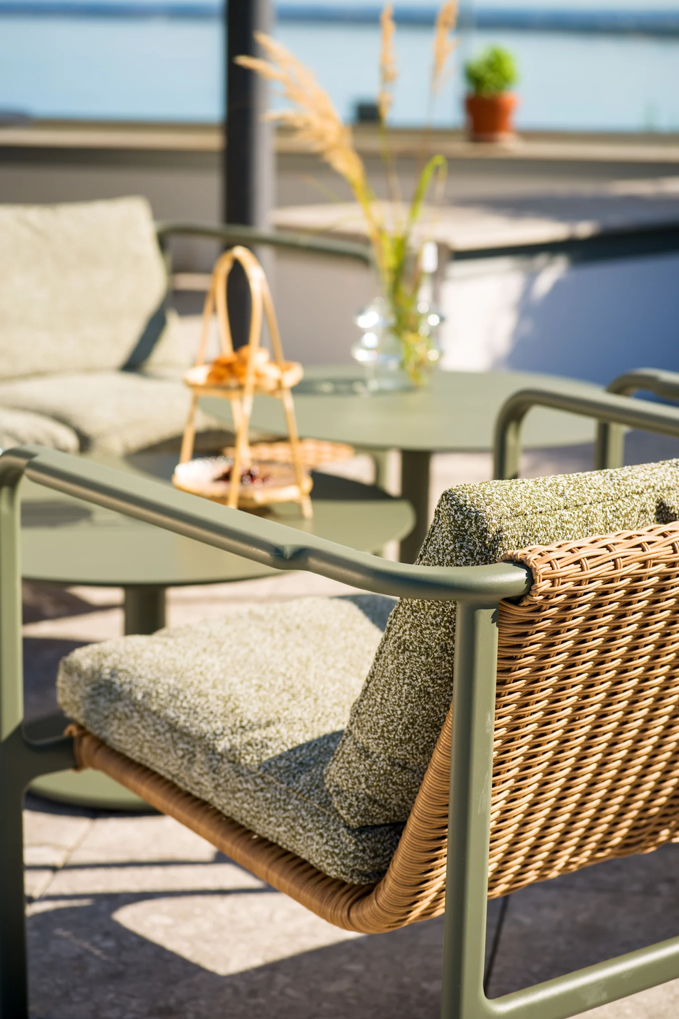 Outdoor-Terrassenstuhl mit olivgrünem Rahmen, geflochtener Rattanlehne und grünen Kissen. Passender Tisch und Stuhl im unscharfen Hintergrund am Wasser.