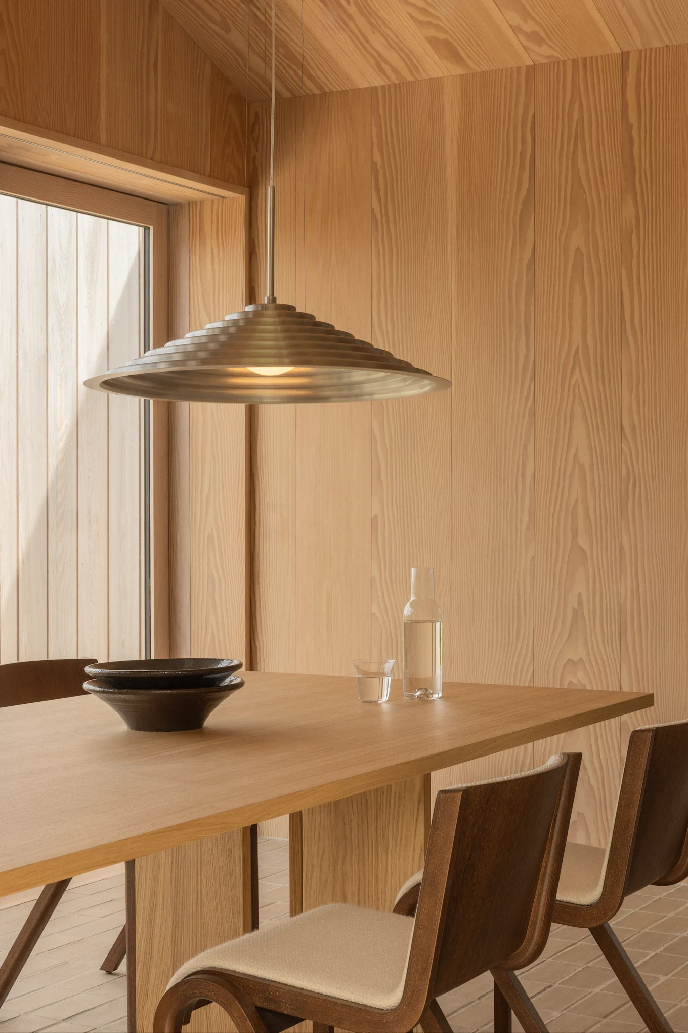 Modernes Esszimmer mit hellen Holzwänden, einem Holztisch mit zwei dunklen Schüsseln, einer Wasserflasche und einer abgestuften Metall-Pendelleuchte.