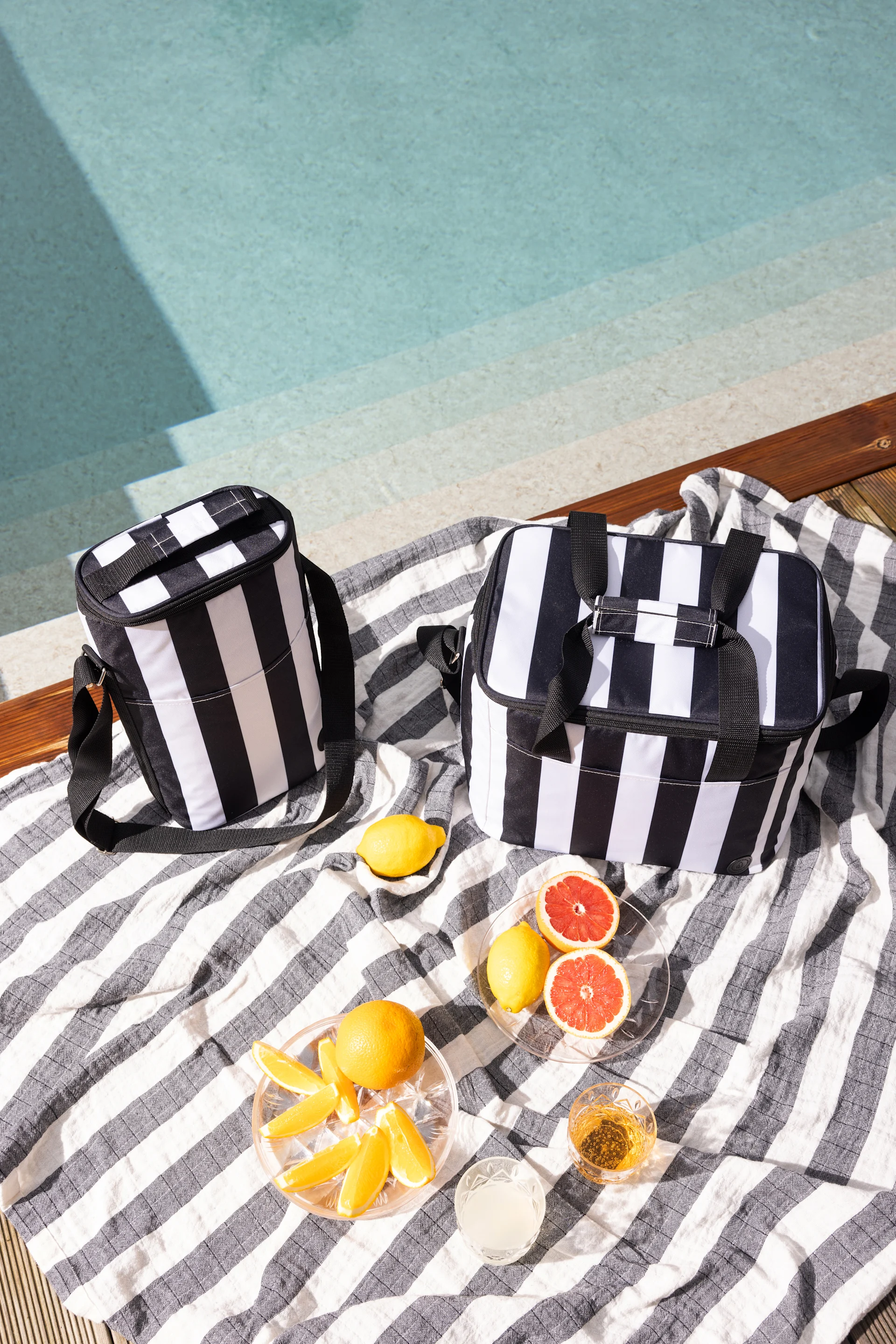 Zwei schwarz-weiß gestreifte Kühltaschen auf einem gestreiften Handtuch neben einem Pool, mit Zitrusfrüchten und Getränken.