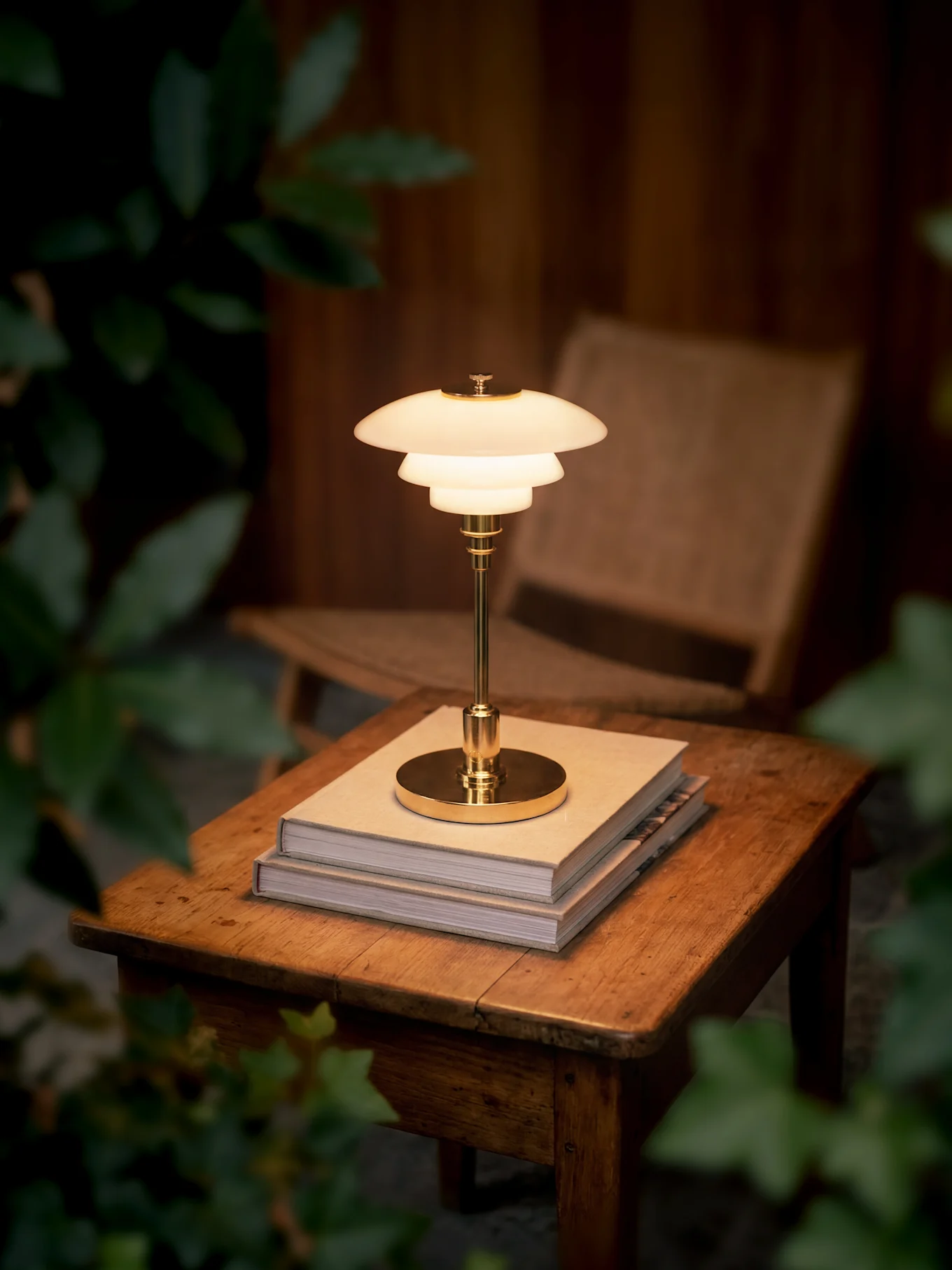 Eine leuchtende, mehrstufige Messing-Tischlampe steht auf zwei Büchern auf einem Holztisch, umgeben von dunklem Laub.
