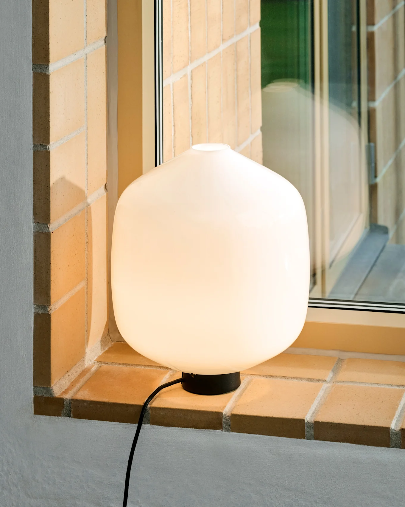 Eine leuchtende, weiße, abgerundete Tischlampe mit schwarzem Sockel und Kabel steht auf einer hellen Ziegel-Fensterbank.