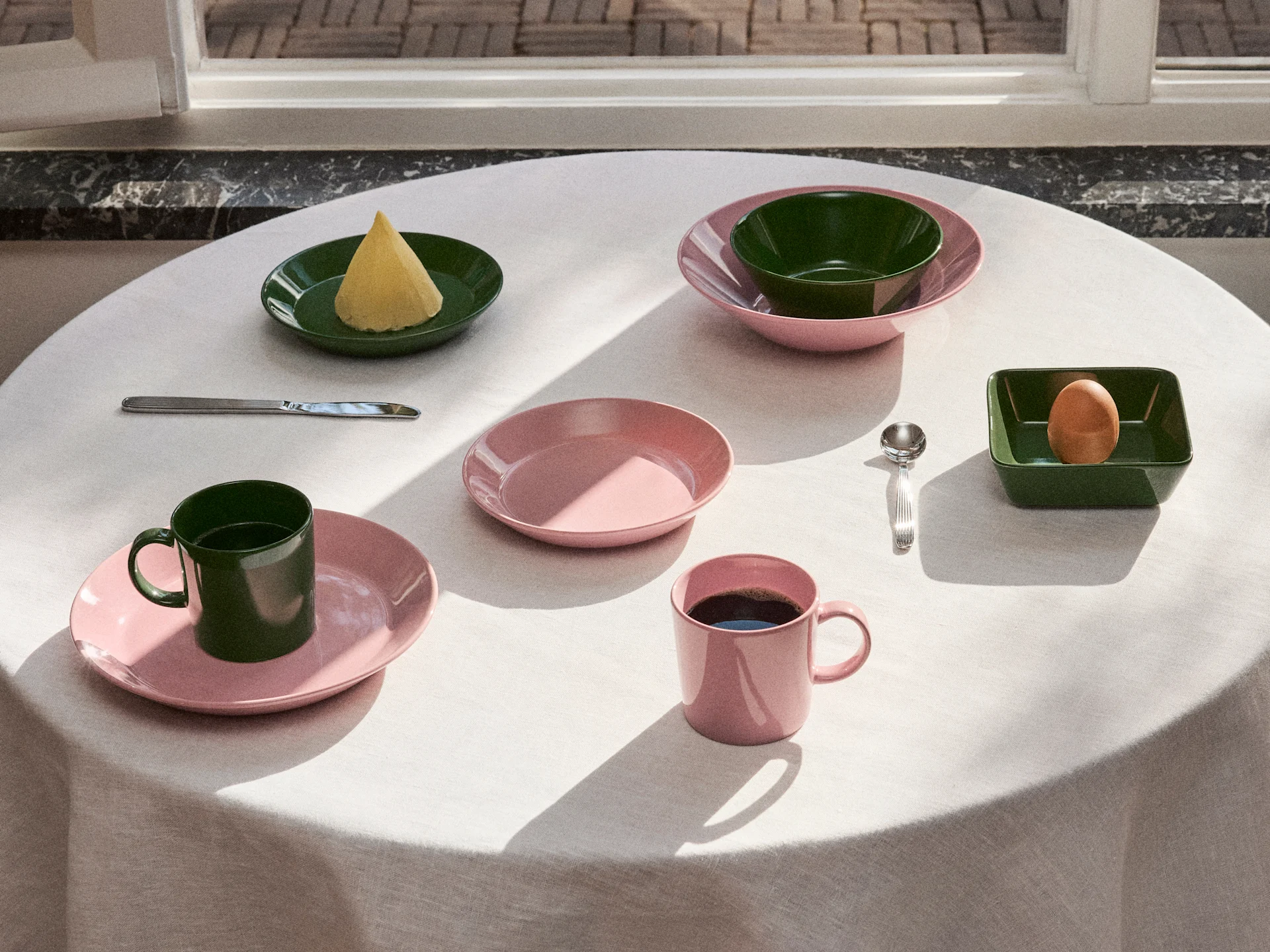 Tisch gedeckt mit rosa und dunkelgrünem Geschirr, Tellern, Schalen und Tassen, mit Butter, einem Ei und Kaffee im Naturlicht.