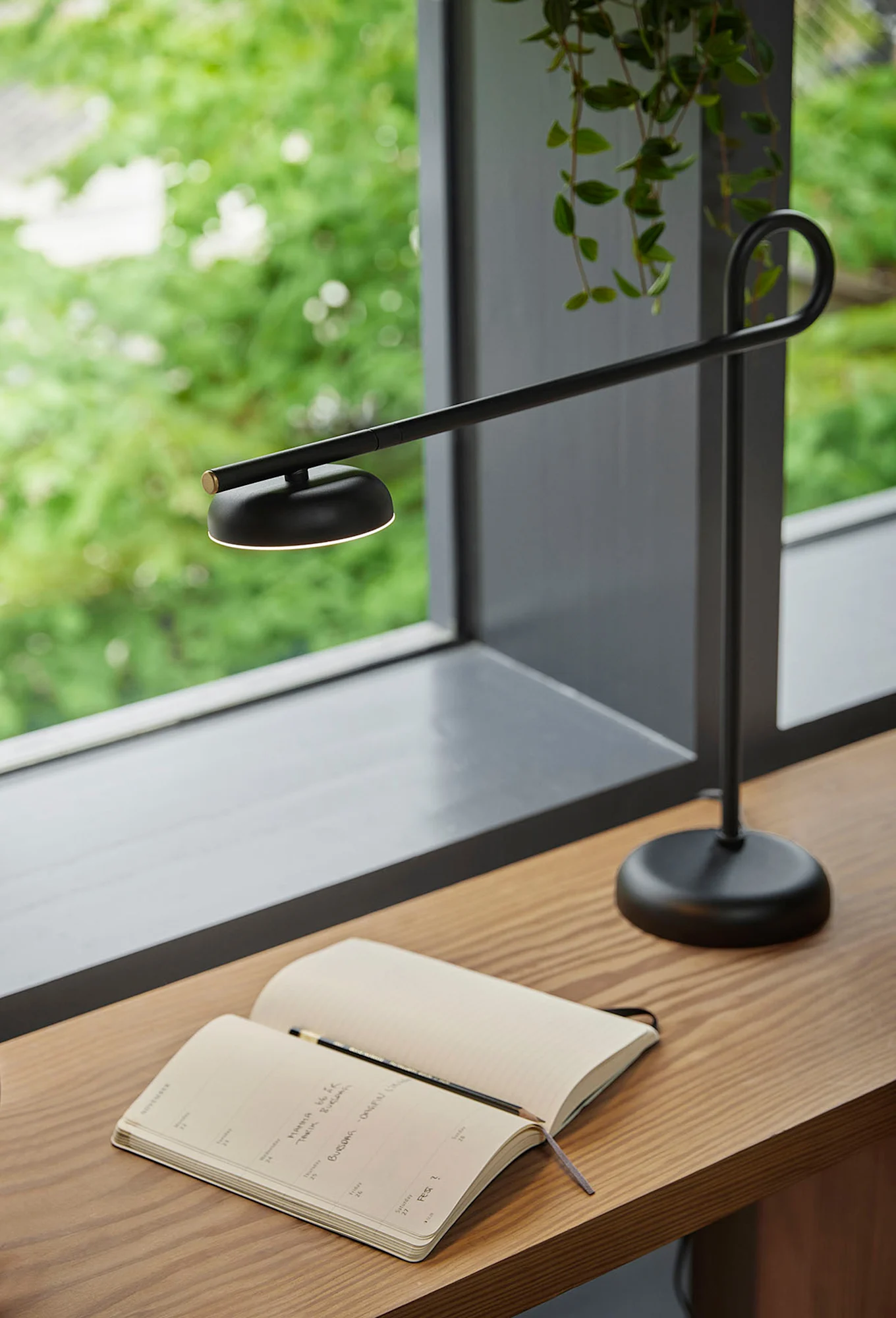 Eine moderne schwarze Schreibtischlampe beleuchtet ein offenes Notizbuch und einen Bleistift auf einem Holzschreibtisch neben einem Fenster mit grünem Laub.