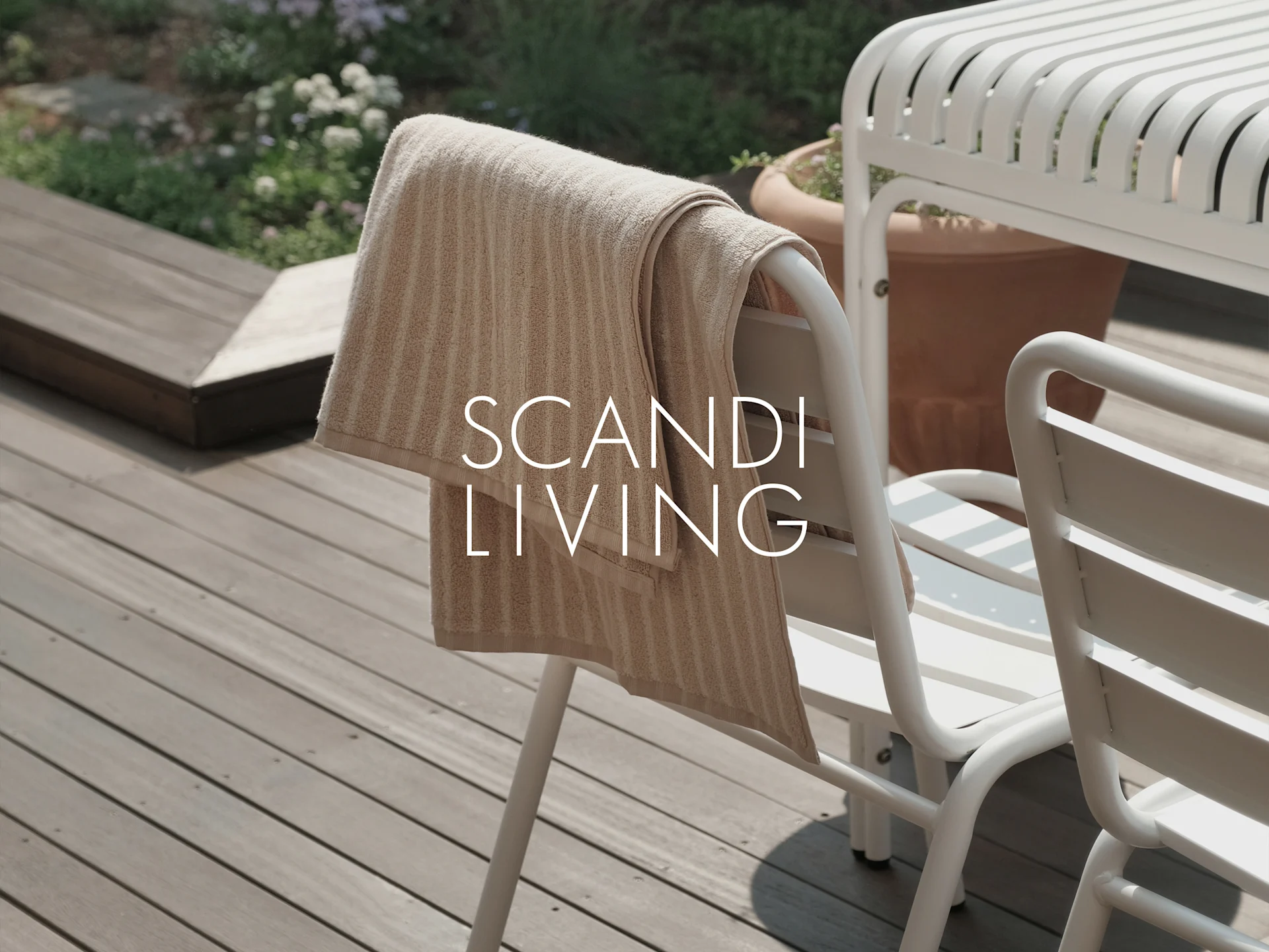 Ein beige gestreiftes Handtuch hängt auf einem weißen Gartenstuhl auf einer Holzterrasse mit Pflanzen. Text: SCANDI LIVING.