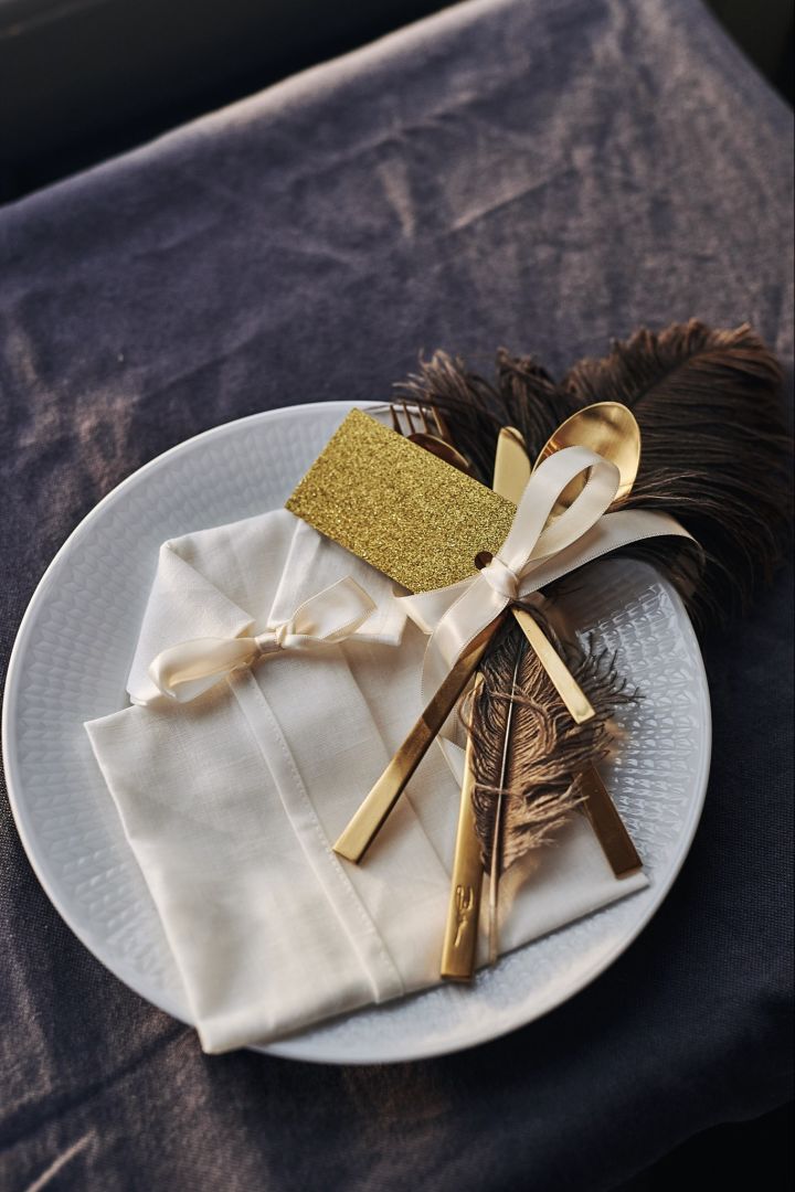 Eine Serviette in Form eines Hemdes liegt auf dem Teller mit Besteck, Tischkarten und einer dekorativen Feder. 