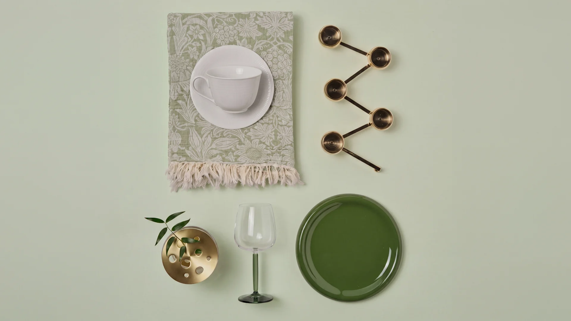 Weiße Teetasse, gemusterte Serviette, Messing-Messlöffel, Messingvase, grünes Weinglas, dunkelgrüner Teller.