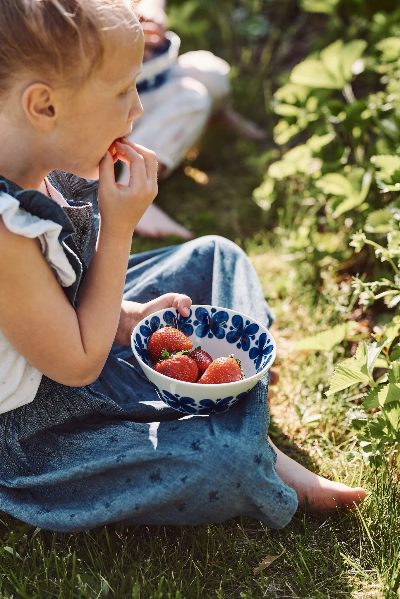 Auch Schweden hat im Sommer viele Erdbeeren zu bieten. Hier sitzt ein Mädchen auf einer Wiese, in der Hand hält sie die Mon Amie-Schale von Rörstrand, welche mit Erdbeeren gefüllt ist.