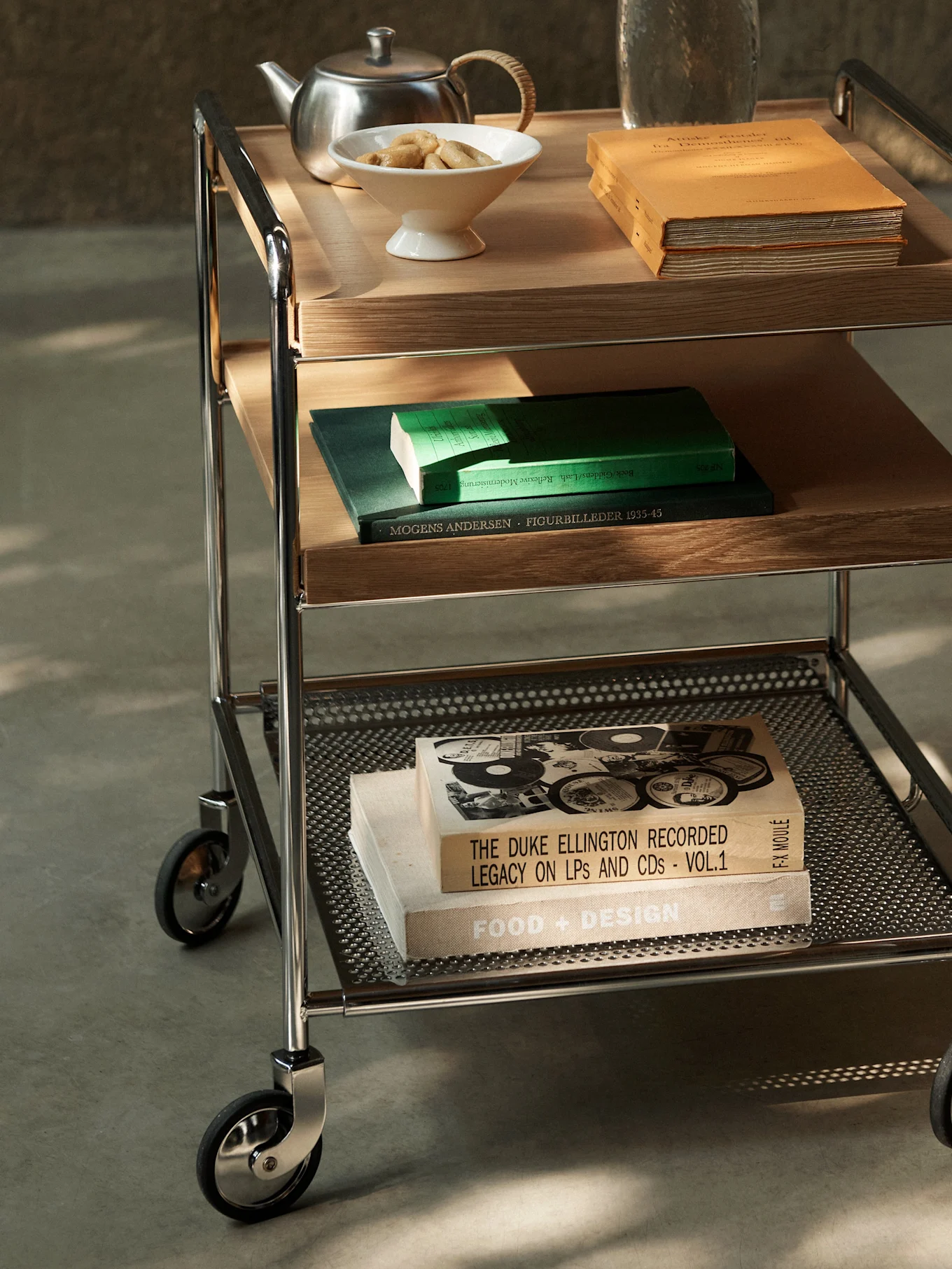 Ein dreistöckiger Servierwagen mit Chromgestell, Holz- und perforierten Metallablagen, mit Büchern, Teekanne und Keksen.