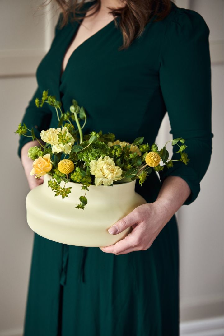  Eine Frau in einem grünen Kleid hält die zartgelbe, niedrige Keramikvase aus der Kollektion Dorotea für Gense. 