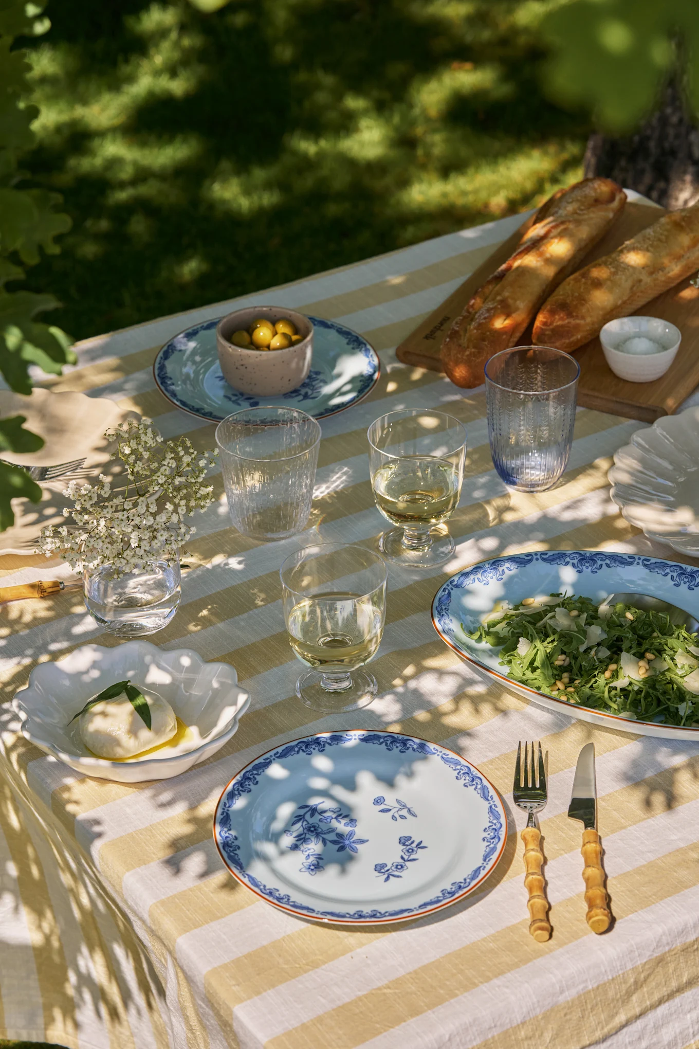 Gedeckter Esstisch im Freien mit weiß-gelb gestreifter Tischdecke, blauen Blumentellern, Wein, Salat, Mozzarella, Oliven und Baguettes.