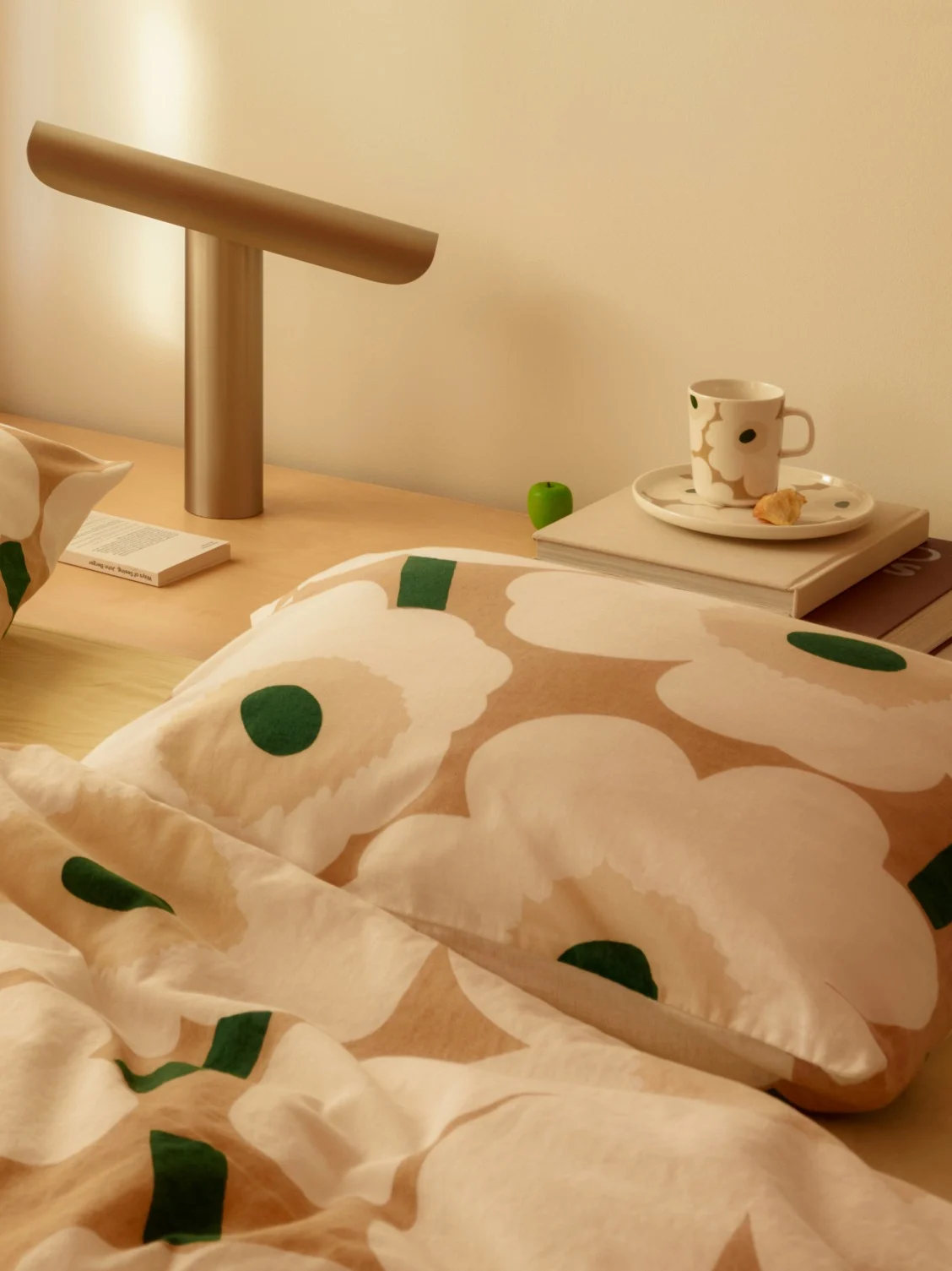 Nachttischszene mit weiß-beiger Blumenbettwäsche, einer modernen Lampe, einer Tasse und einem Buch auf einem Holztisch.