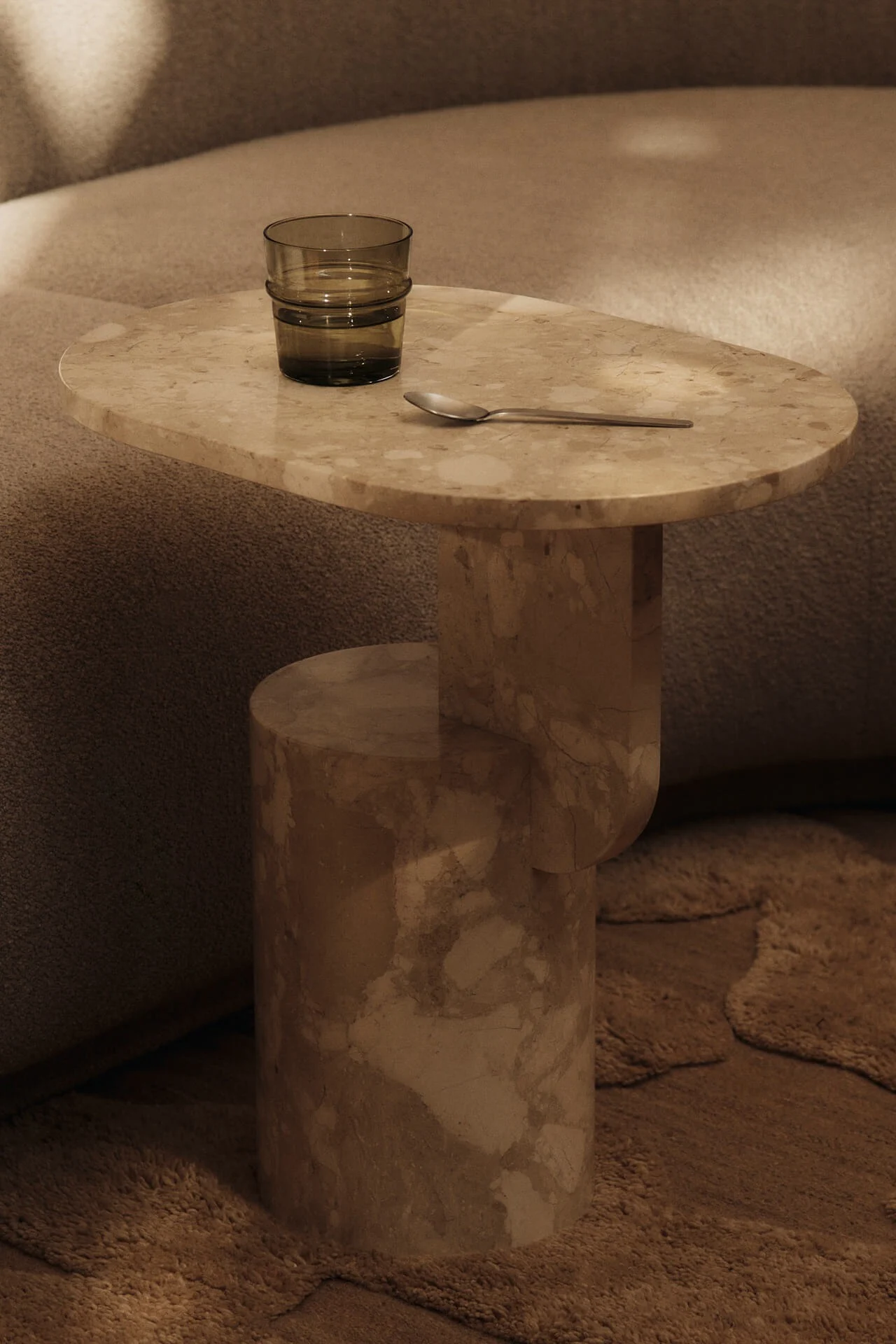 Beiger Marmor-Beistelltisch mit einem dunkelgrünen Wasserglas und einem Silberlöffel, neben einem strukturierten Sofa und Teppich.