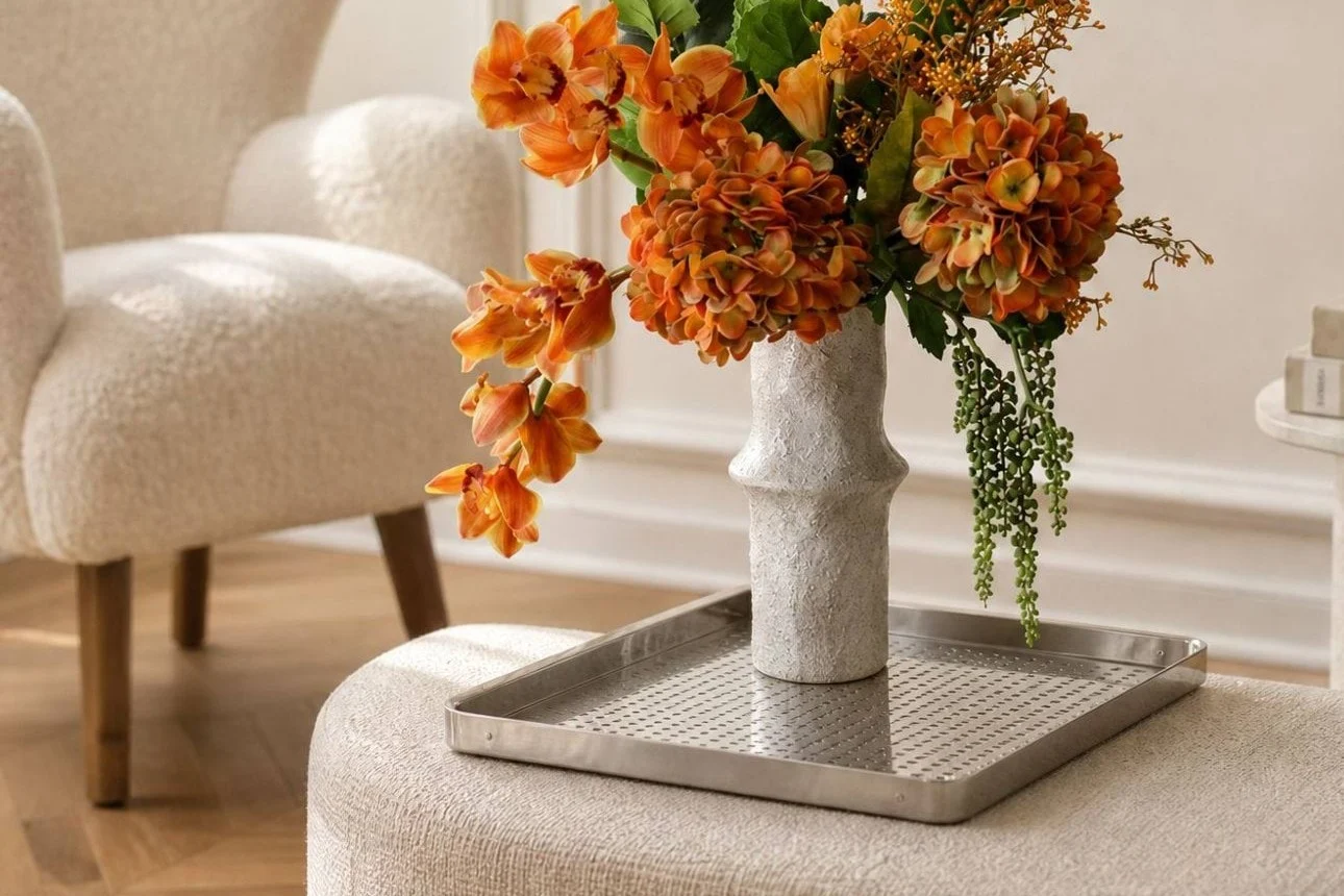 Leuchtend orangefarbene Blumen in einer strukturierten weißen Vase auf einem silbernen Tablett, platziert auf einem hellen Hocker in einem modernen Wohnzimmer.