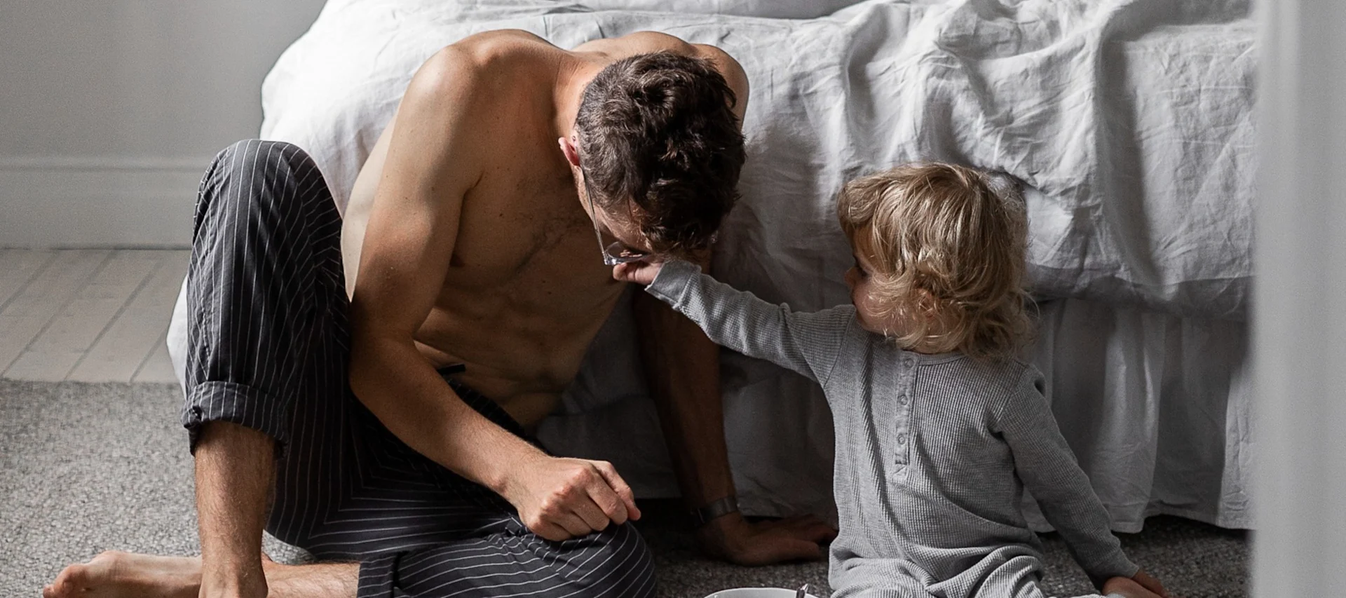 Geschenke zum Vatertag: Hier sehen Sie einen Mann mit einem Kleinkind vor einem Bett sitzen.