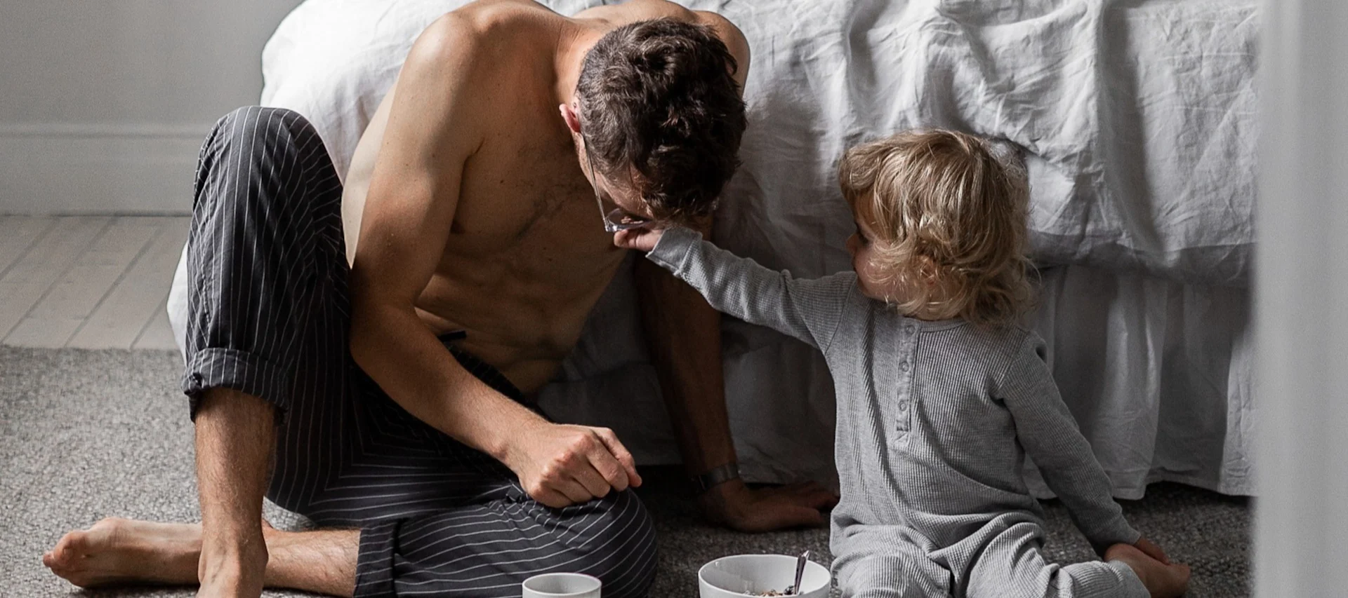 Vater und Kind sitzen im Schlafzimmer auf dem Boden, Kind berührt spielerisch Vaters Brille, Frühstück in der Nähe.