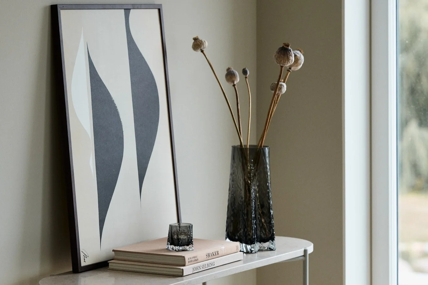 Hier sehen Sie die Gry Vase 30 cm und ein Gry Windlicht, beides in der Farbe Smoke von Cooee Design zusammen mit einem Poster auf einem Beistelltisch stehen. 