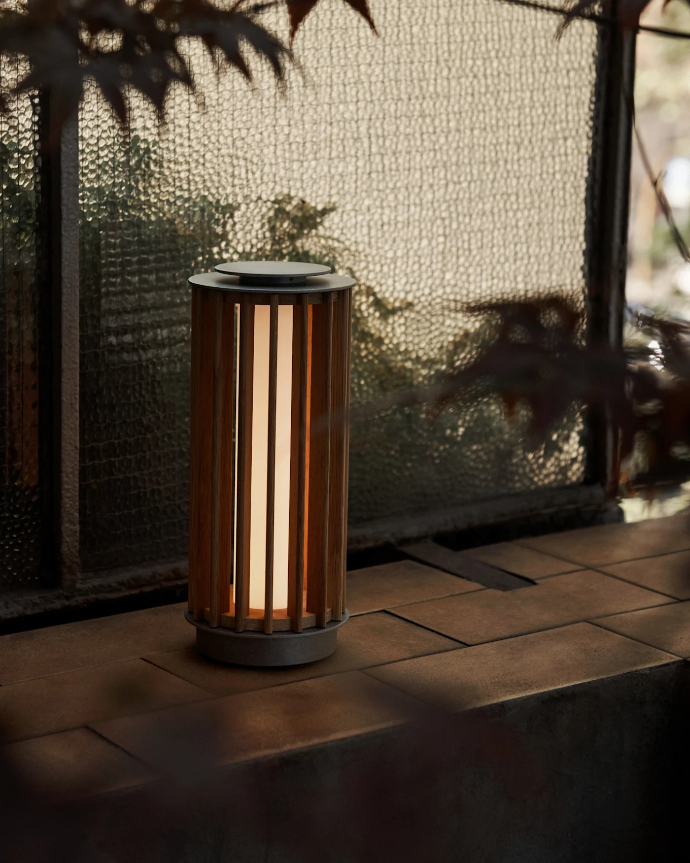Eine zylindrische tragbare Lampe mit einem Lattenholzgehäuse und warmem Licht, auf einer gefliesten Fläche an einem strukturierten Glasfenster.