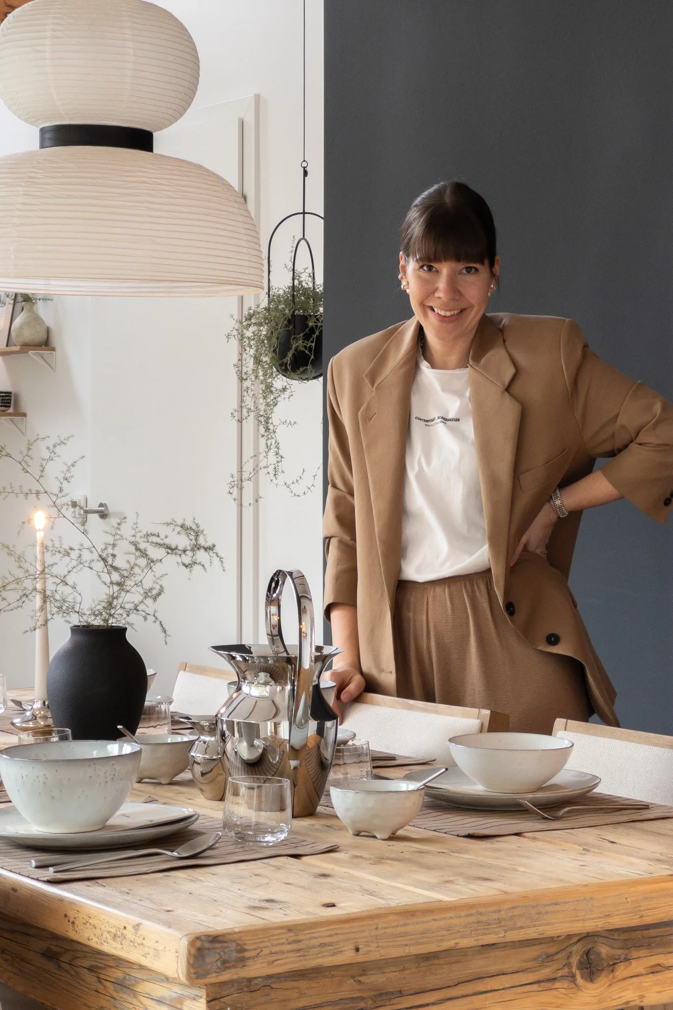 Eine lächelnde Frau in braunem Anzug steht hinter einem rustikalen Holztisch, gedeckt mit weißem Geschirr, silbernen Kannen und einer Kerze.
