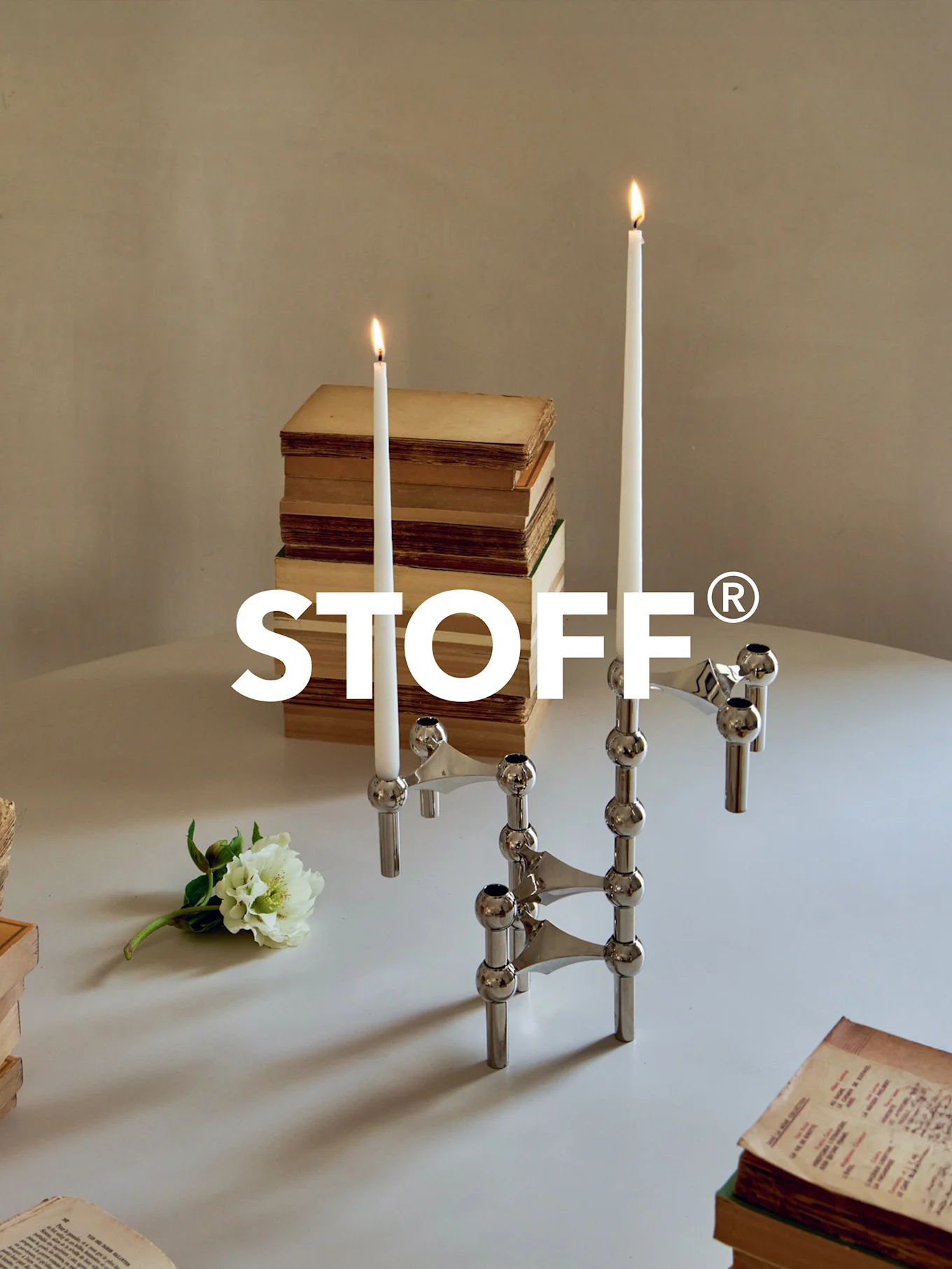Polierter Chrom-Modulkerzenständer mit zwei brennenden Kerzen, umgeben von alten Büchern und einer weißen Blume auf einem Tisch. STOFF® Logo.