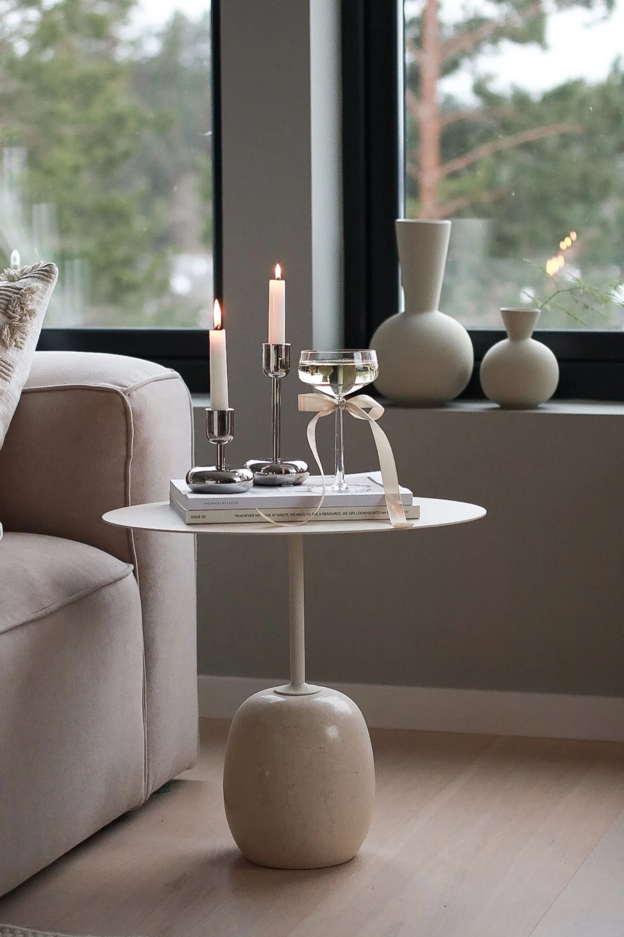 Eine gemütliche Wohnzimmerszene mit brennenden Kerzen, Büchern und einem Champagnerglas auf einem weißen Beistelltisch neben einem beigen Sofa, mit Vasen auf der Fensterbank.