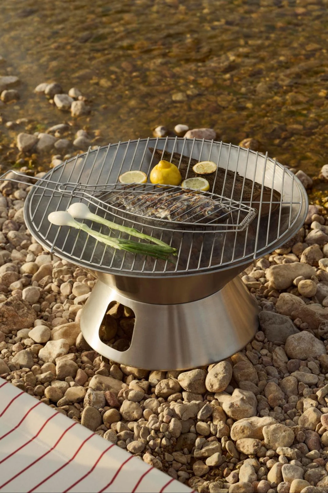 Ein tragbarer Edelstahlgrill mit einem ganzen Fisch in einem Grillkorb, Zitronen und Frühlingszwiebeln an einem felsigen Flussufer.