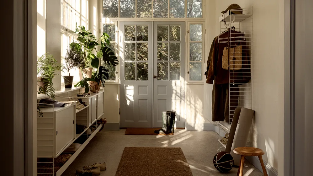 Heller Eingangsbereich mit weißen Modulregalen, natürlichem Licht, Pflanzen, Schuhen, Kleidung, einem Hut und doppelten Glastüren.