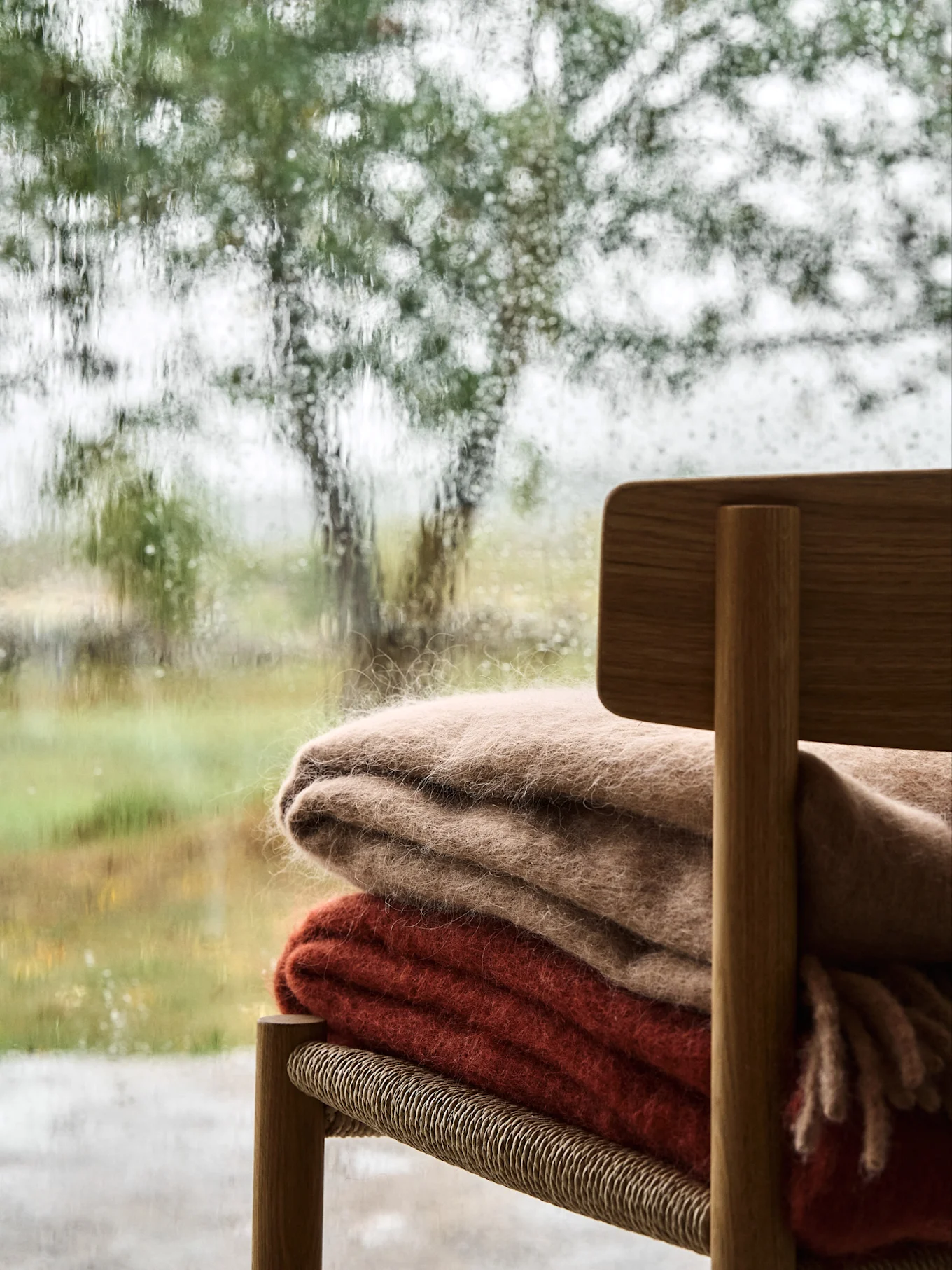 Gemütliche Szene mit zwei flauschigen Decken auf einem Holzstuhl, gesehen durch ein regenstreifiges Fenster mit Blick auf eine verschwommene grüne Landschaft.