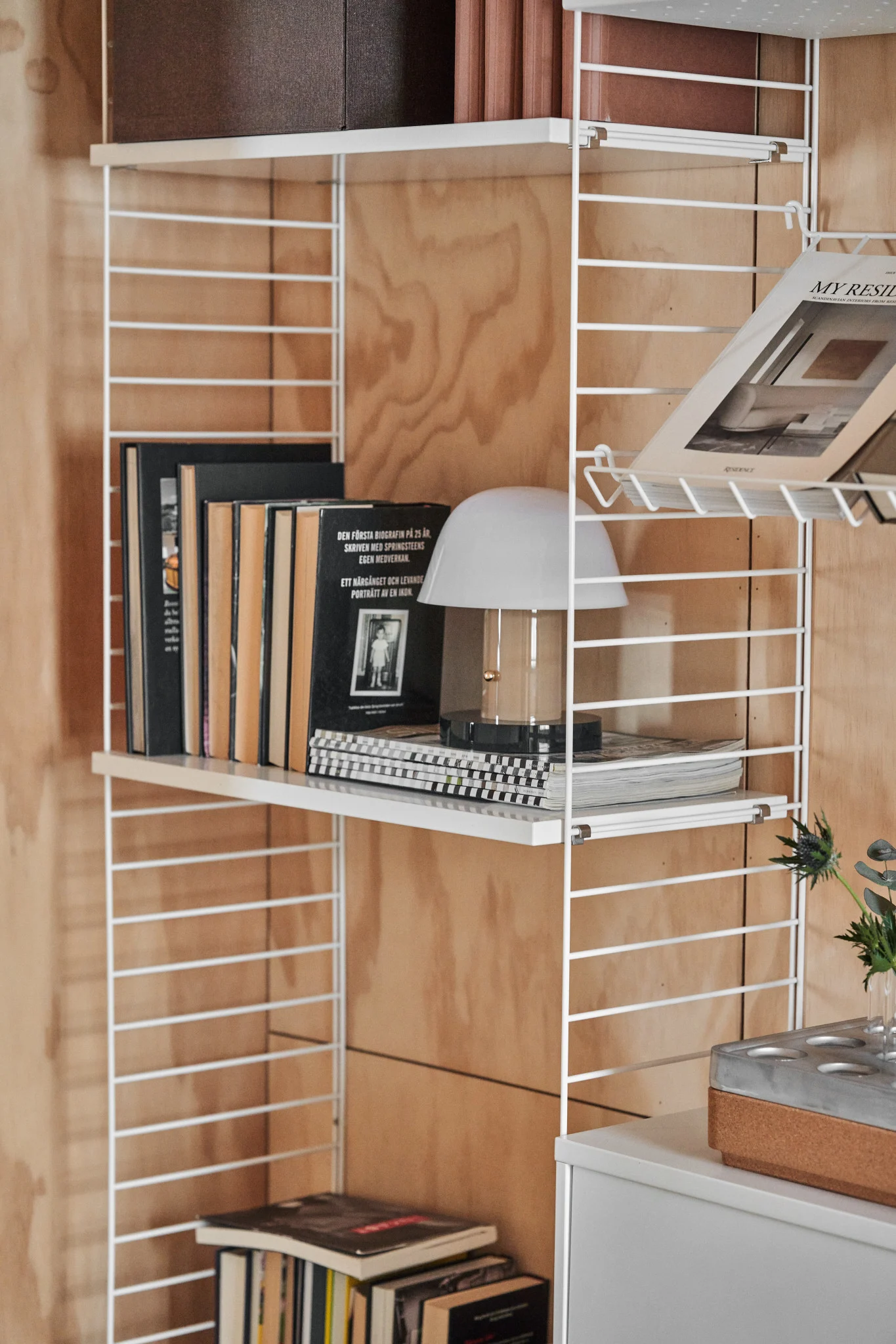 Weißes Drahtregal an einer Holzwand, mit Büchern, einer weißen Tischlampe und einem Magazin. Ein grauer Schrank steht darunter.
