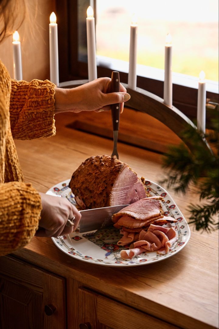 Schwedische Weihnachten: Eine Frau schneidet einen traditionellen nordischen Weihnachtsschinken in einem Fenster neben einem Adventskerzenhalter auf. 