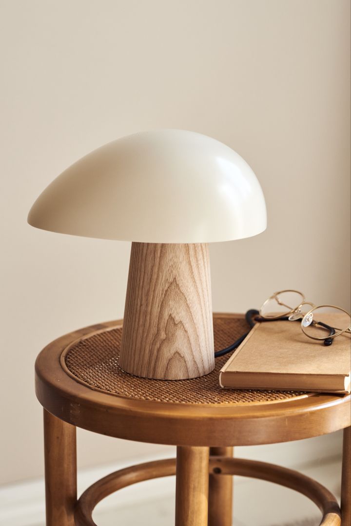 Beleben Sie Ihr Zuhause mit einer modernen Tischleuchte – Hier sehen Sie die Fritz Hansen Night Owl Tischleuchte in Weiß und hellem Holz auf einem Rattan-Hocker.