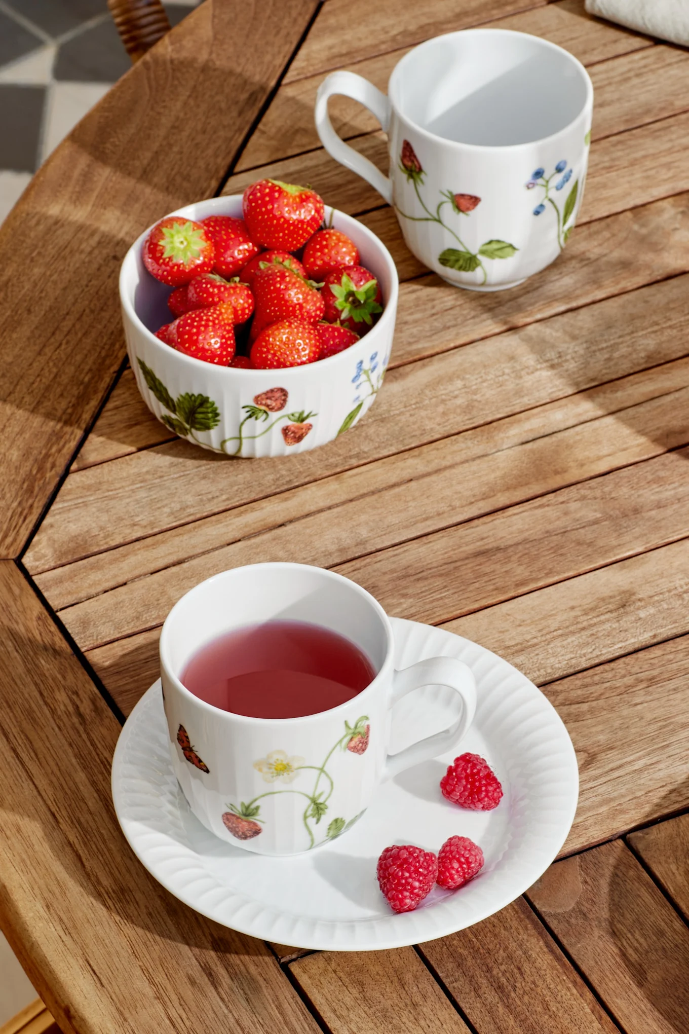 Eine weiße Tasse mit rosa Tee und Untertasse mit Himbeeren, eine Schale Erdbeeren und eine leere Tasse auf einem Holzlamellentisch im Sonnenlicht.