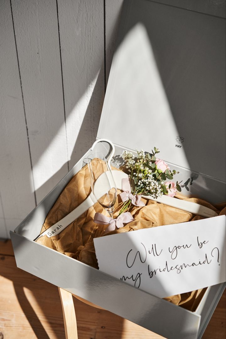 Lassen Sie sich von unseren einzigartigen Hochzeitsideen inspirieren, wie z. B. einem personalisierten Brautjungfernantrag in einer grauen Aufbewahrungsbox von HAY, mit einem personalisierten Kleiderbügel für das Kleid und einem Sektglas zur Feier des Tages. 