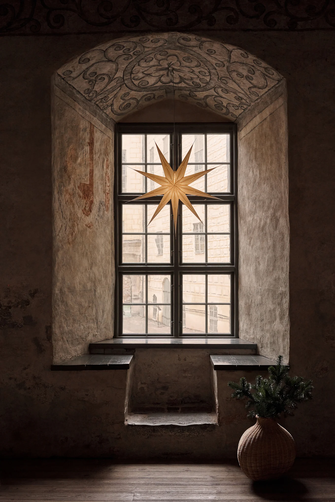 Ein altes Rundbogenfenster mit einem großen Papierstern. Eine Weidenvase mit Kiefernzweigen steht rechts auf dem Boden.