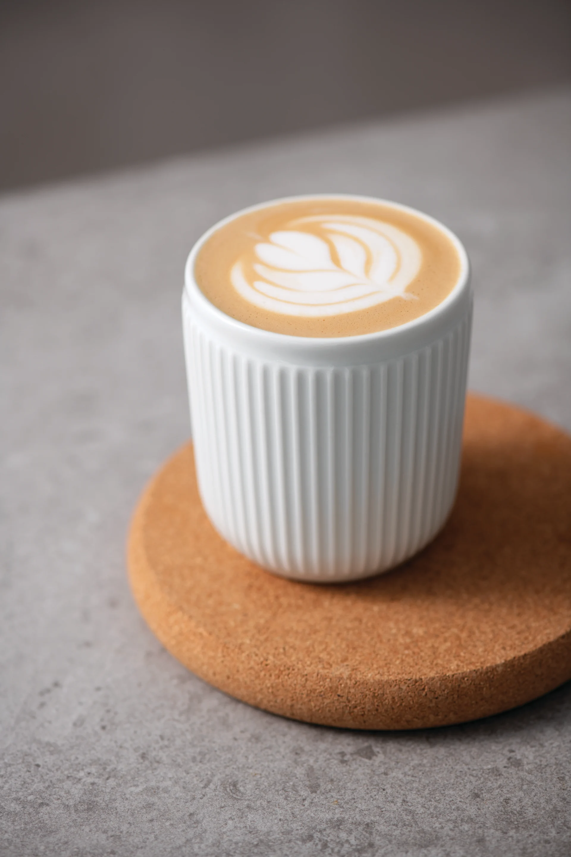 Eine weiße gerippte Tasse Latte mit kunstvoller Blattkunst steht auf einem runden Korkuntersetzer auf einer grauen Oberfläche.