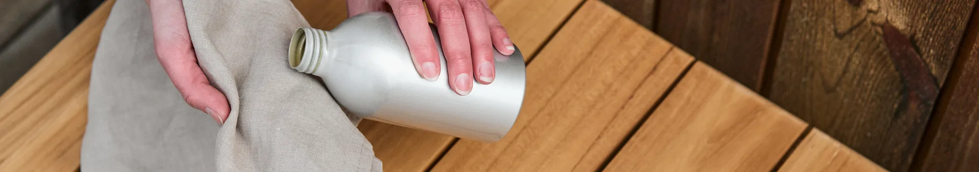 Ein Paar Hände hält ein Tuch und eine Flasche Teaköl bereit, um eine Teakholzbank der Marke 1898 bei Nordic Nest zu ölen. 