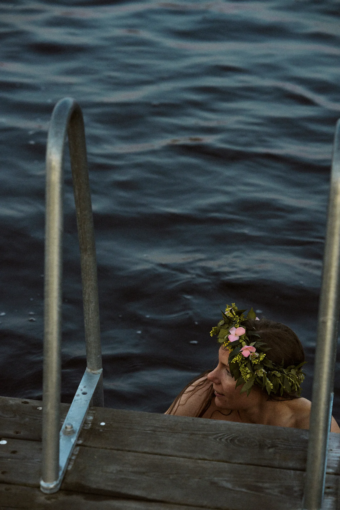 Frau in dunklem Wasser mit Blumenkranz, neben einem Holzsteg mit Metallgeländern.