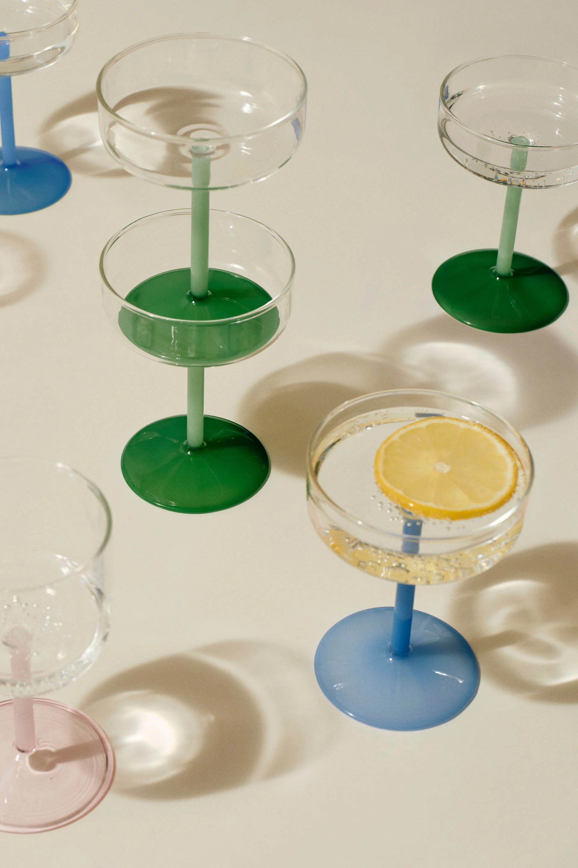 Auswahl an Coupe-Gläsern mit farbigen Stielen und Füßen. Ein Glas enthält eine Zitronenscheibe in sprudelndem Wasser.