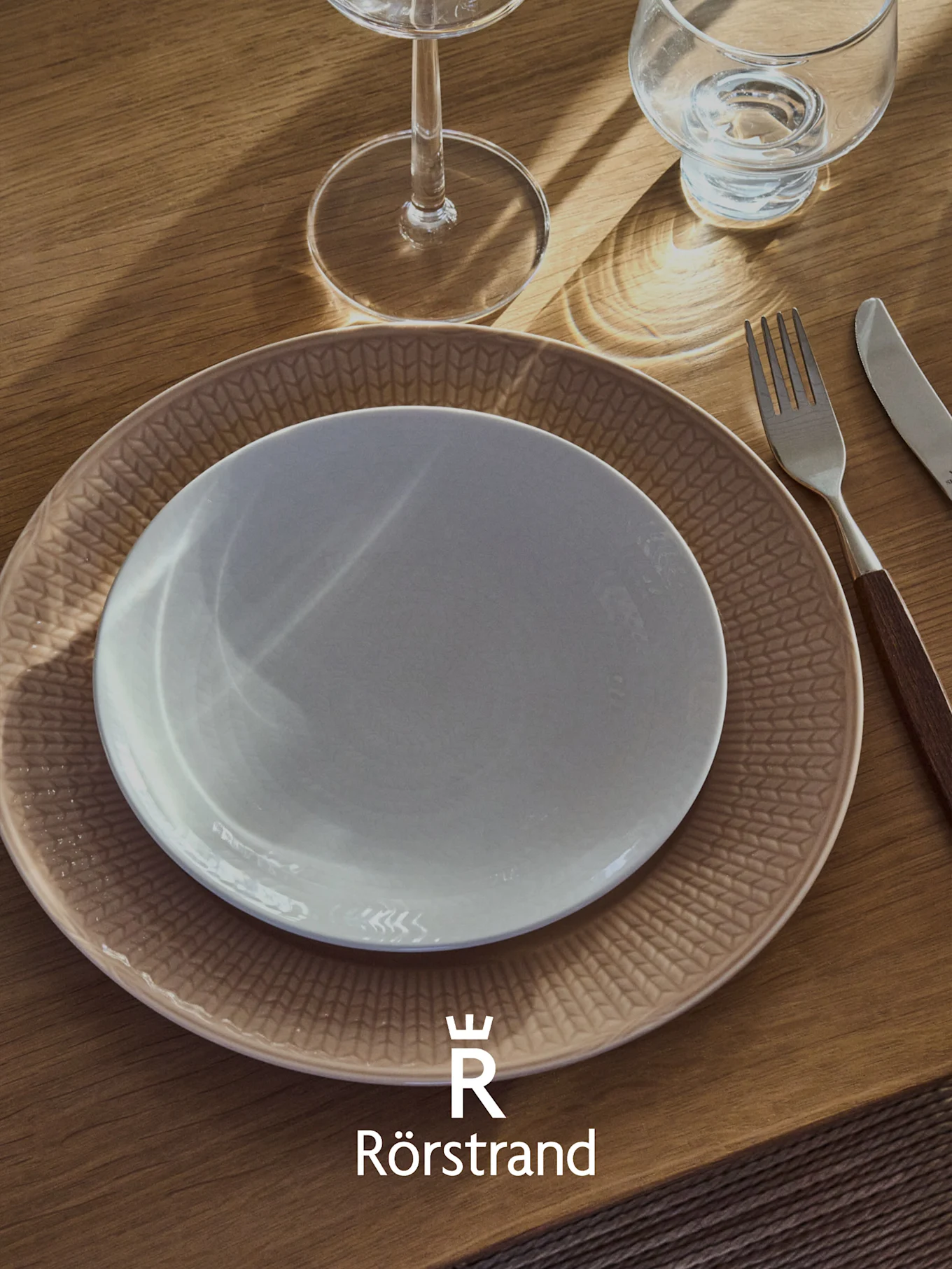 Ein Gedeck mit einem weißen Teller auf einem gemusterten beigen Teller, einer Gabel, einem Messer und zwei Gläsern auf einem Holztisch. Rörstrand-Logo.