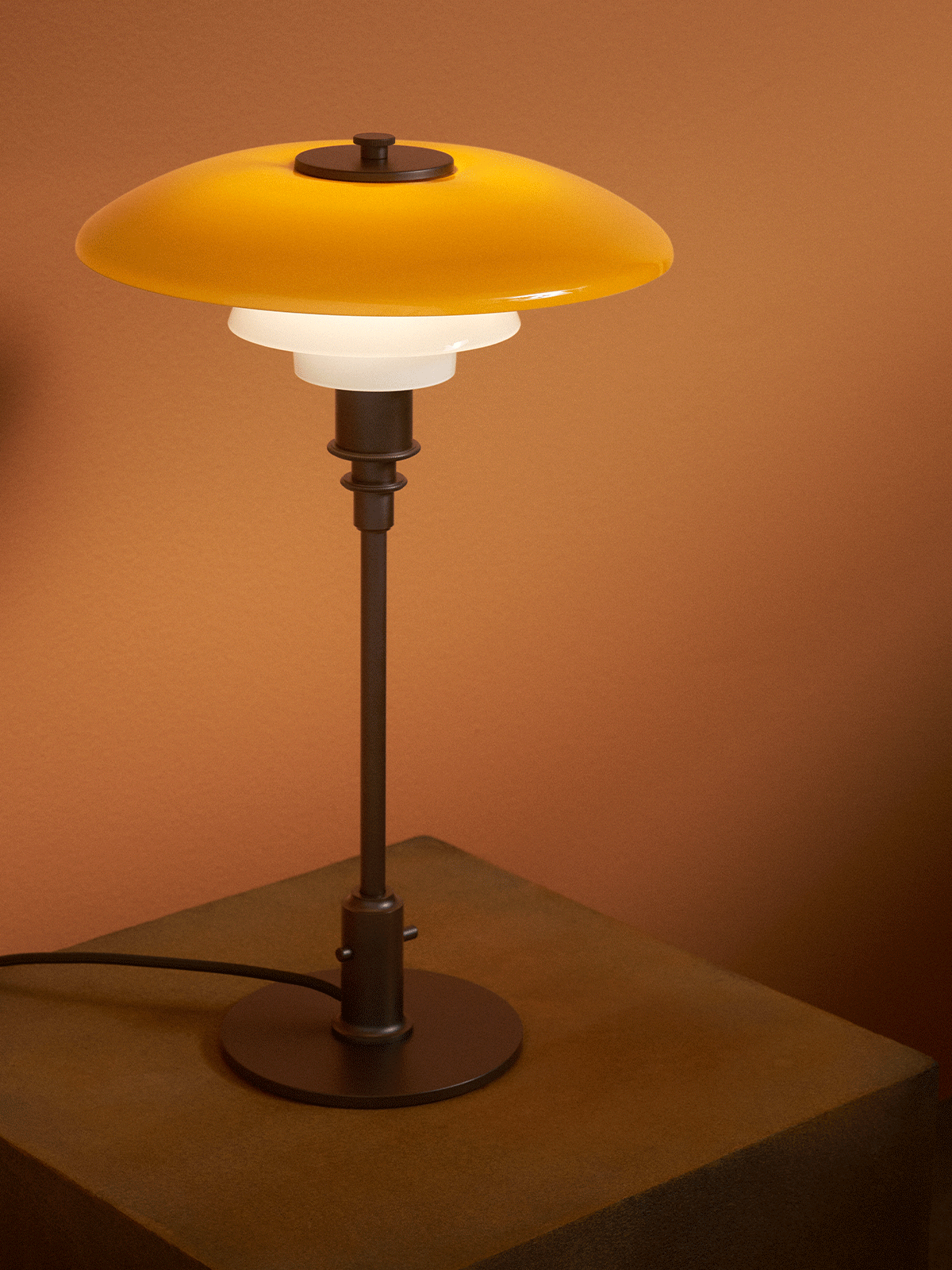 Eine moderne Tischlampe mit gelbem Oberschirm und zwei weißen Schirmen auf einem strukturierten Holzsockel vor brauner Wand.