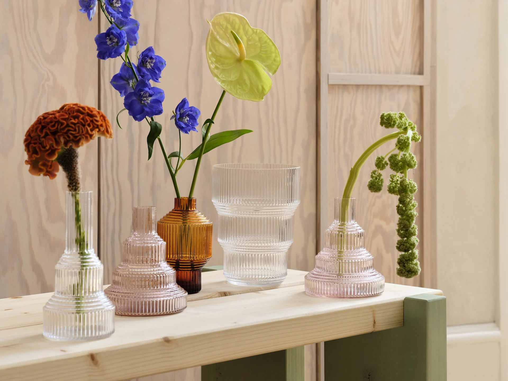 Sortiment gerippter Glasvasen in Klar, Rosa und Bernstein mit verschiedenen Blumen auf einem hellen Holztisch.